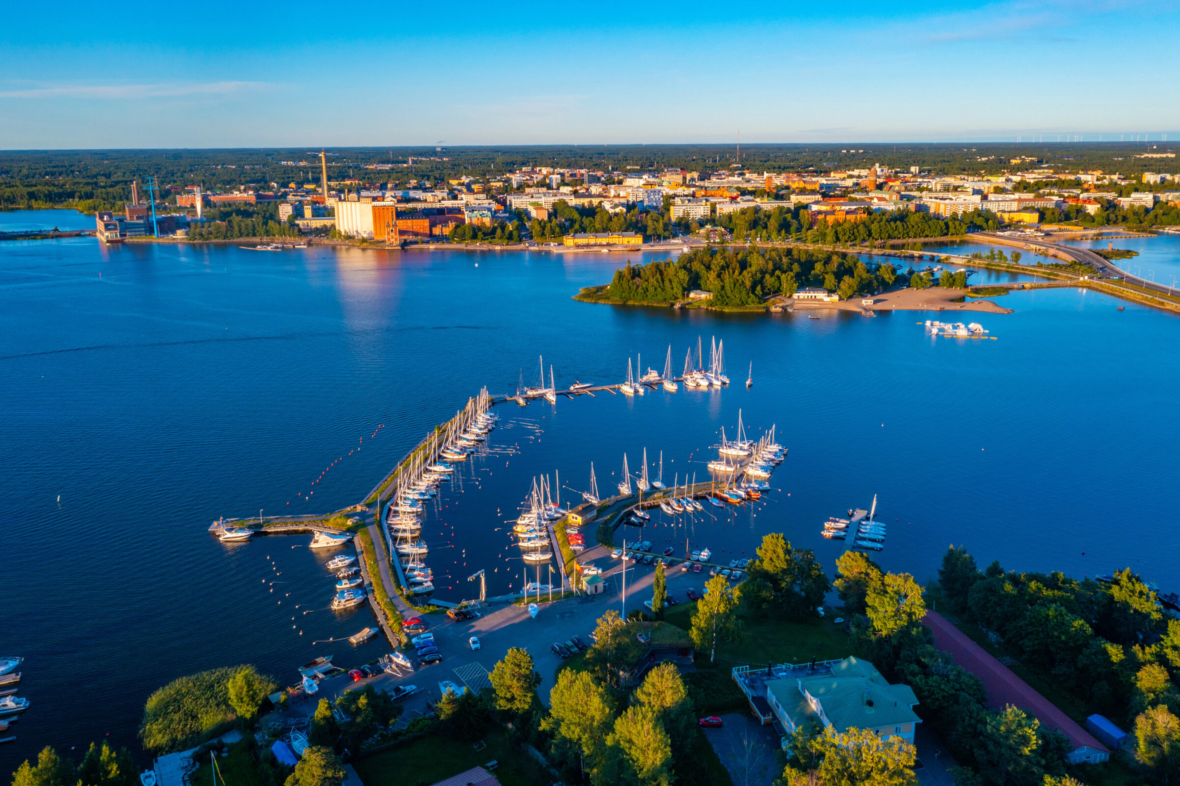 Flygbild av en hamnstad med en småbåtshamn nära stranden omgiven av grönområden. I bakgrunden syns stadens silhuett och industribyggnader.