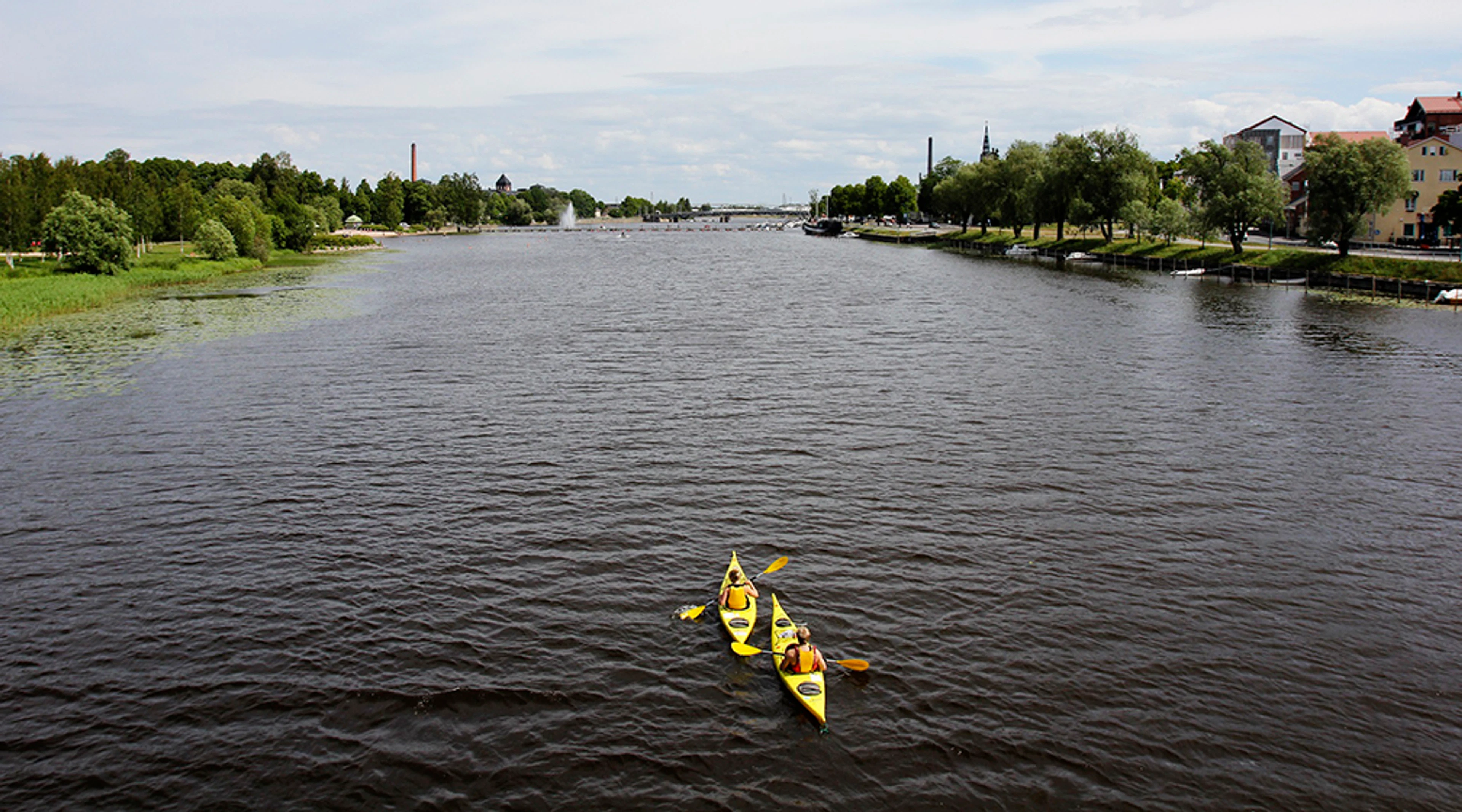Two-person yellow kayak on a wide river, green trees and buildings along the shore.