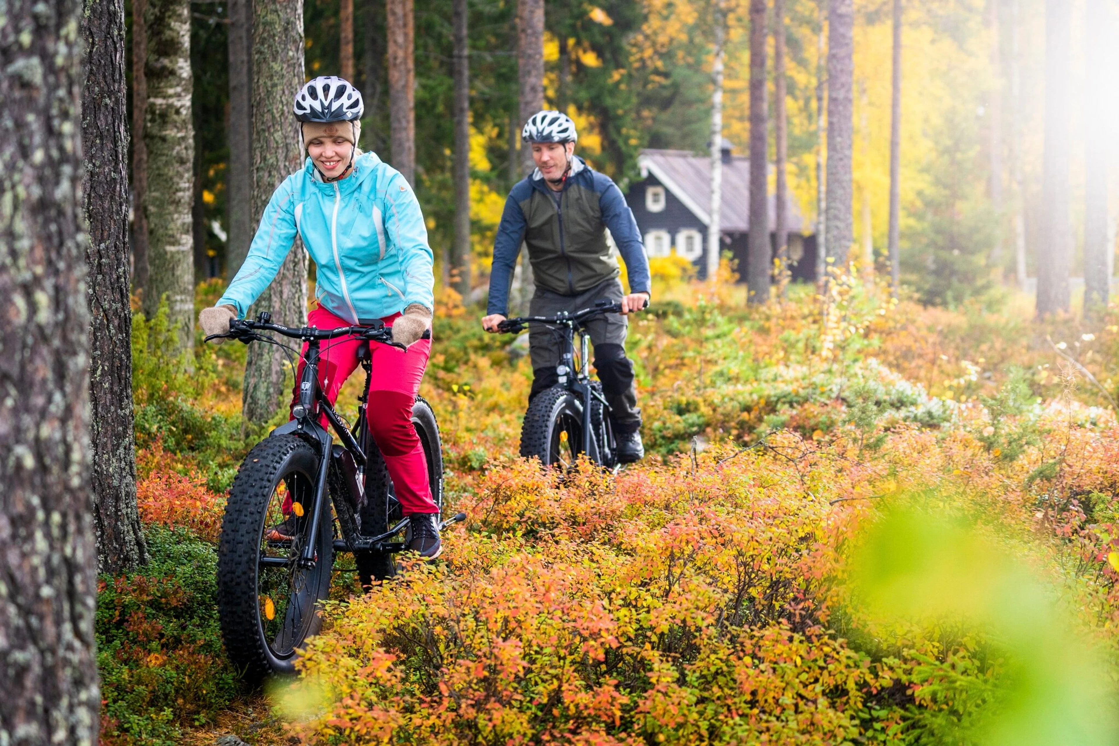 Two cyclists ride fat bikes along a narrow path in an autumn forest. A dark log cabin is in the background.