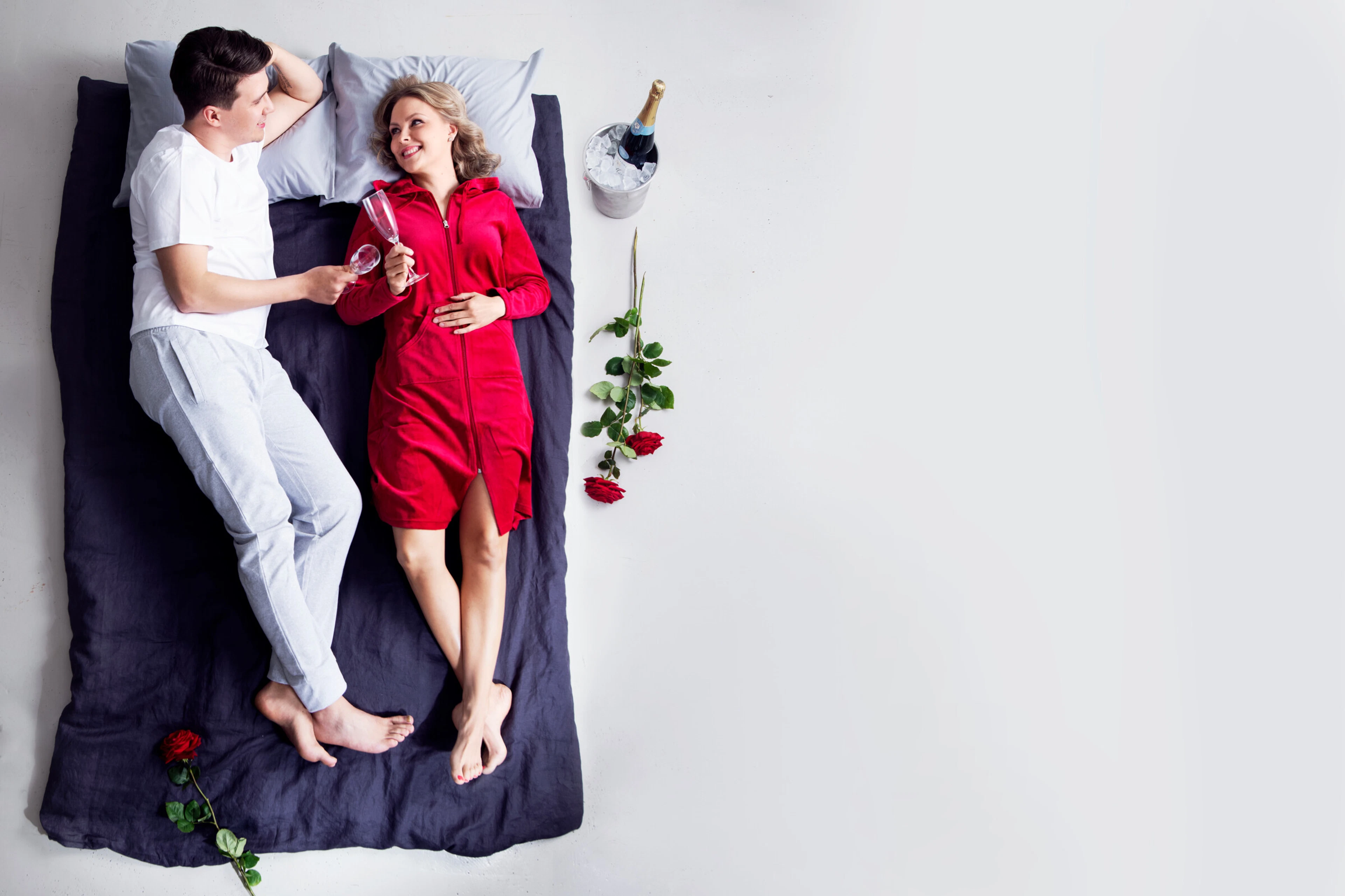 Pari sängyllä, toinen punaisessa asussa ja toinen valkoisessa, pitävät samppanjalaseja. Vieressä jäähdytin ja punaisia ruusuja.