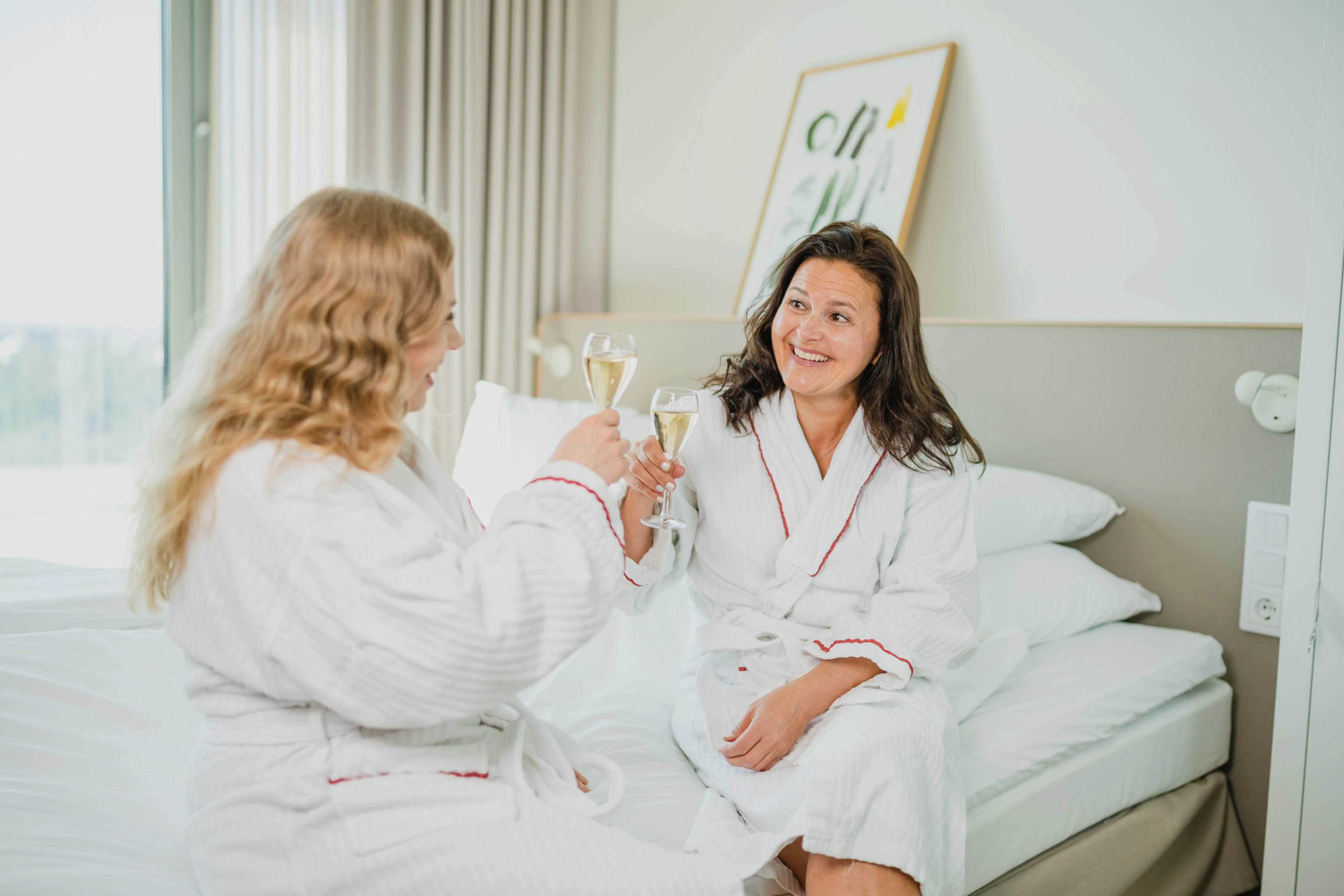 Two people in white bathrobes toast with champagne flutes while sitting on a hotel room bed. Bright decor and large window.