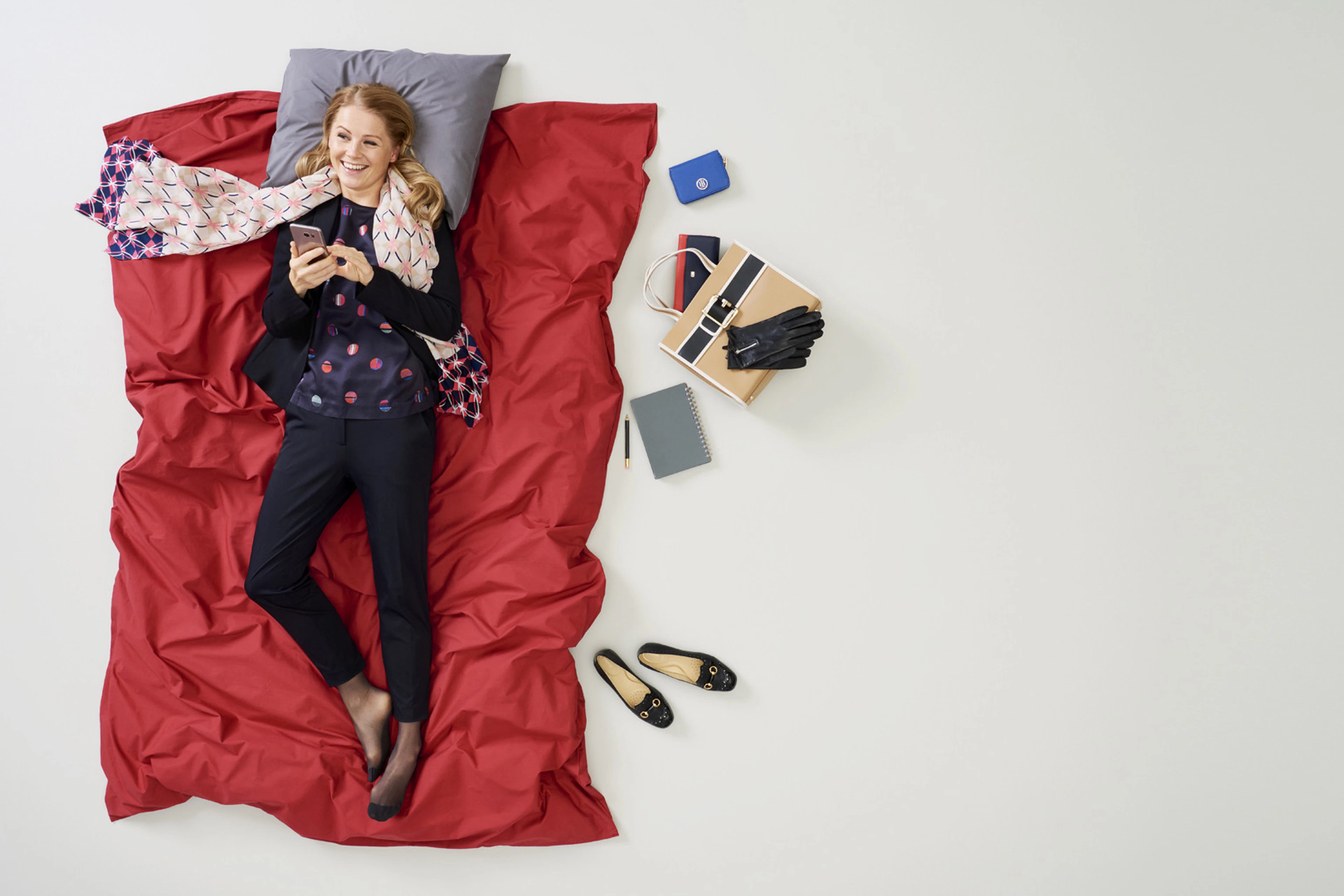 Woman lying on red bedding in dark outfit, looking at phone. Accessories, handbag, and notebook arranged beside her on white background.