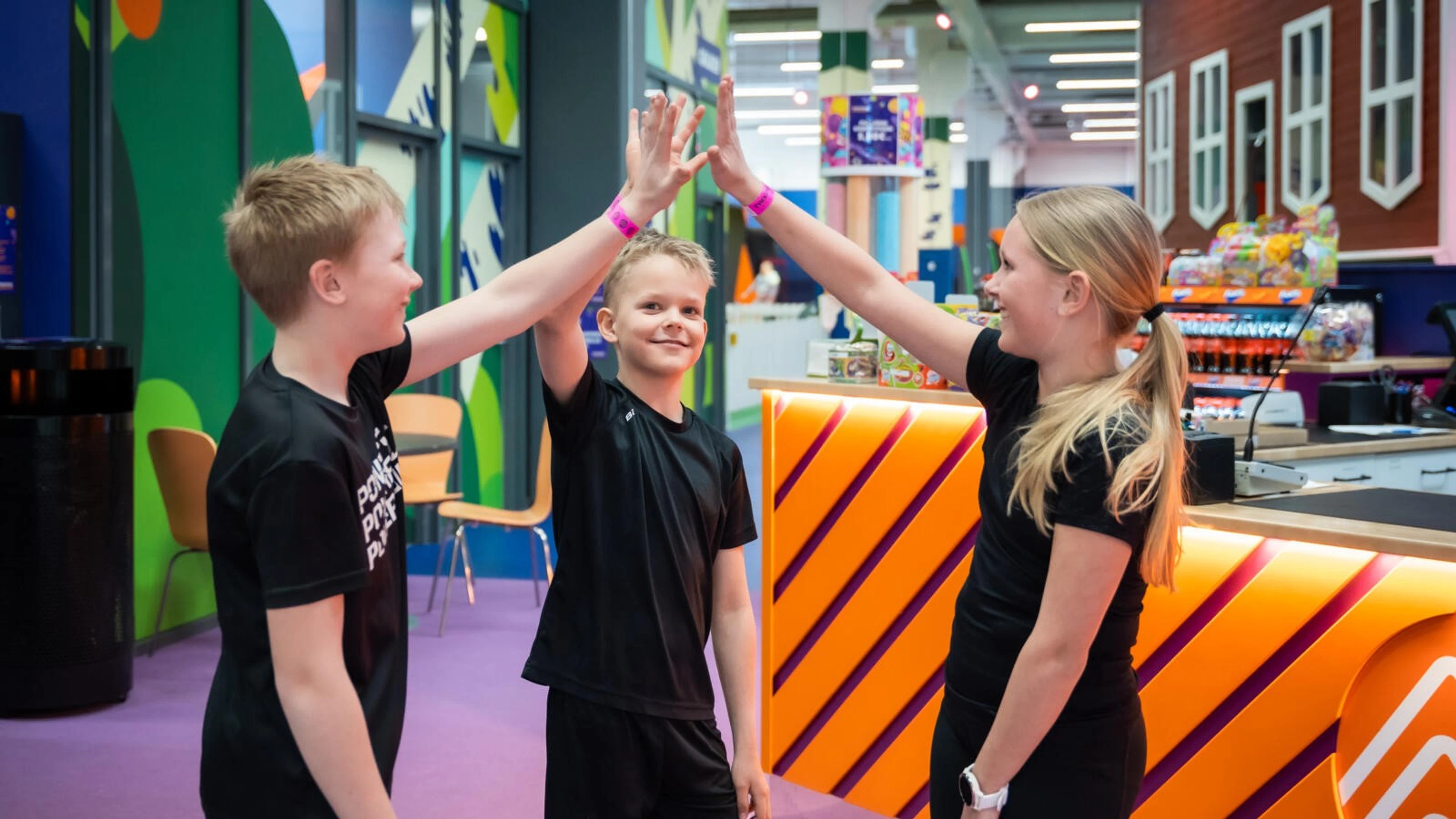 Tre barn ger high five i en färgglad inomhusmiljö med jämnt golv och ljus belysning.