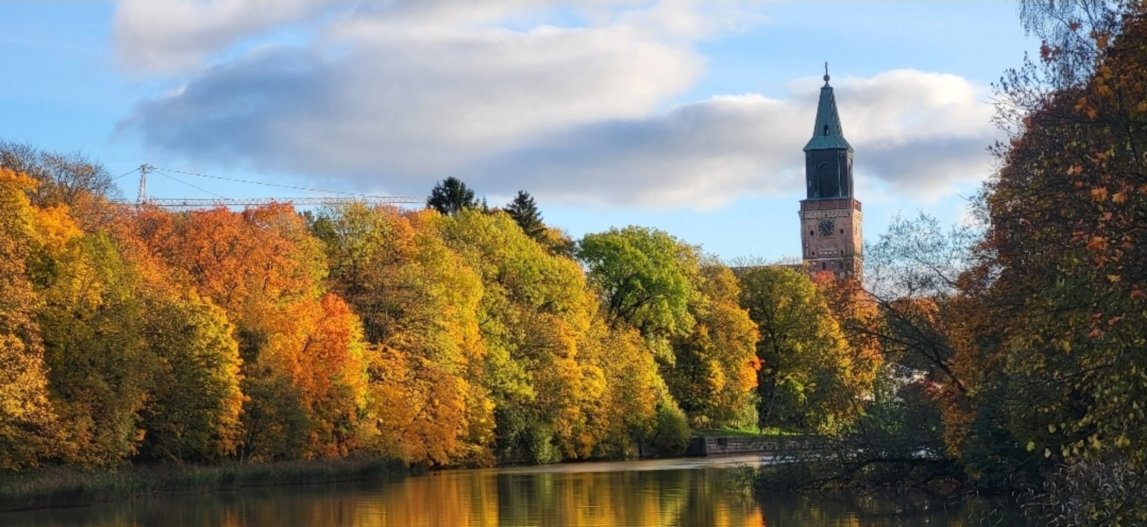 Turku Cathedral rises behind trees in autumn colors, with trees and clouds reflecting in the calm river.