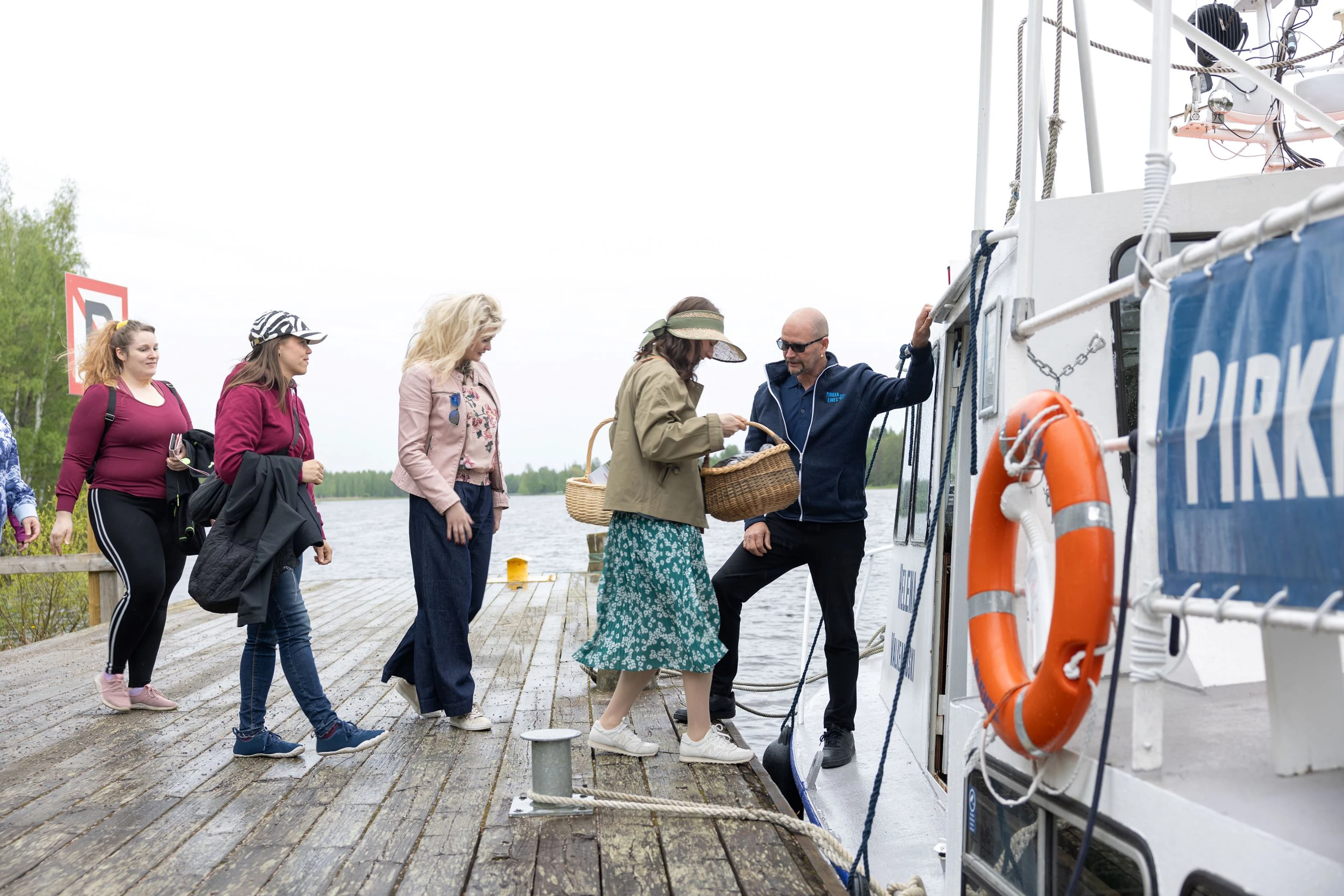 Ryhmä ihmisiä nousemassa veneeseen puulaiturilla. Yhdellä naisella on korikassi. Veneessä on oranssi pelastusrengas.