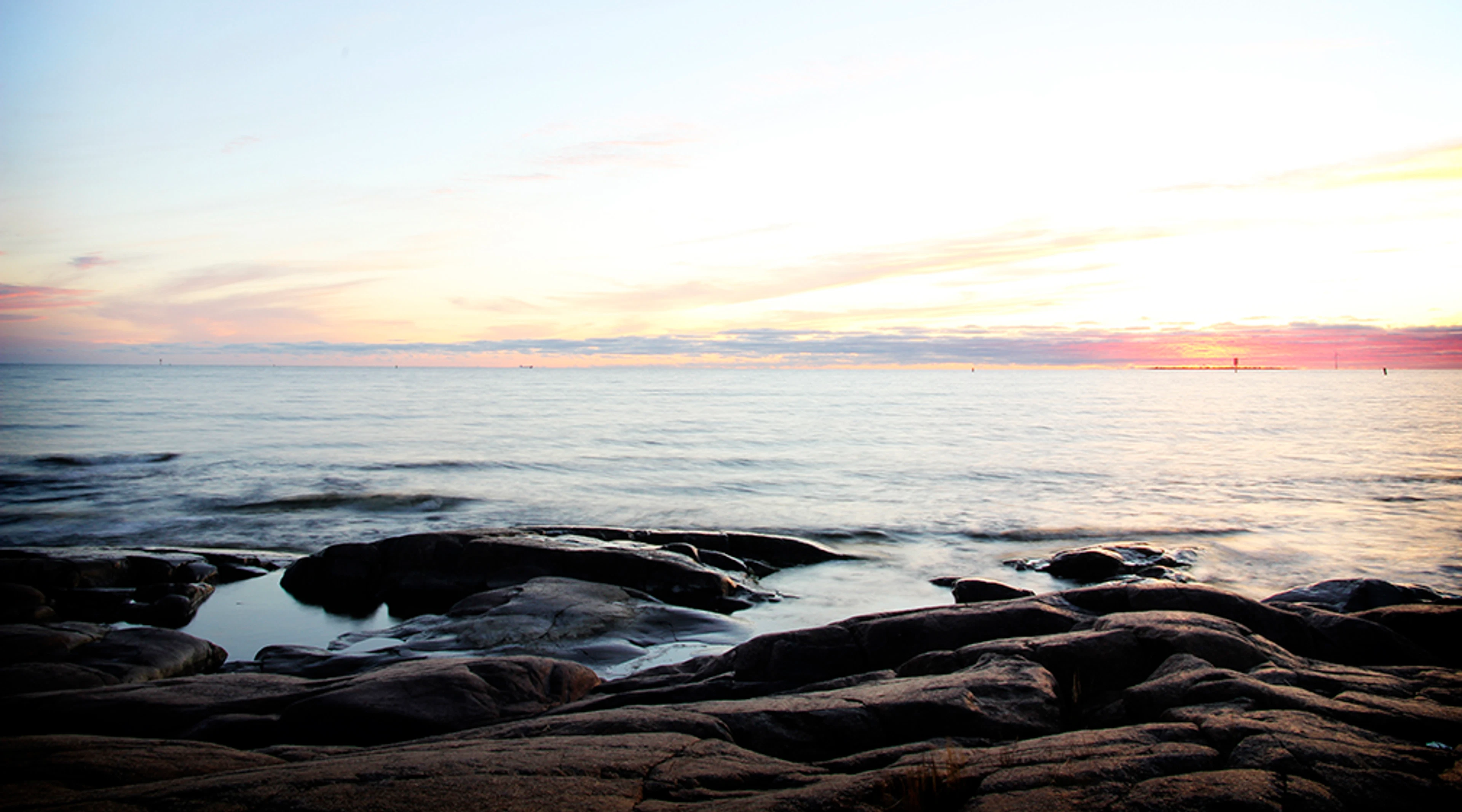 Rauhallinen merimaisema kallioiselta rannalta auringonlaskun aikaan, taivas värjäytynyt pastellisävyisiin pilviin.