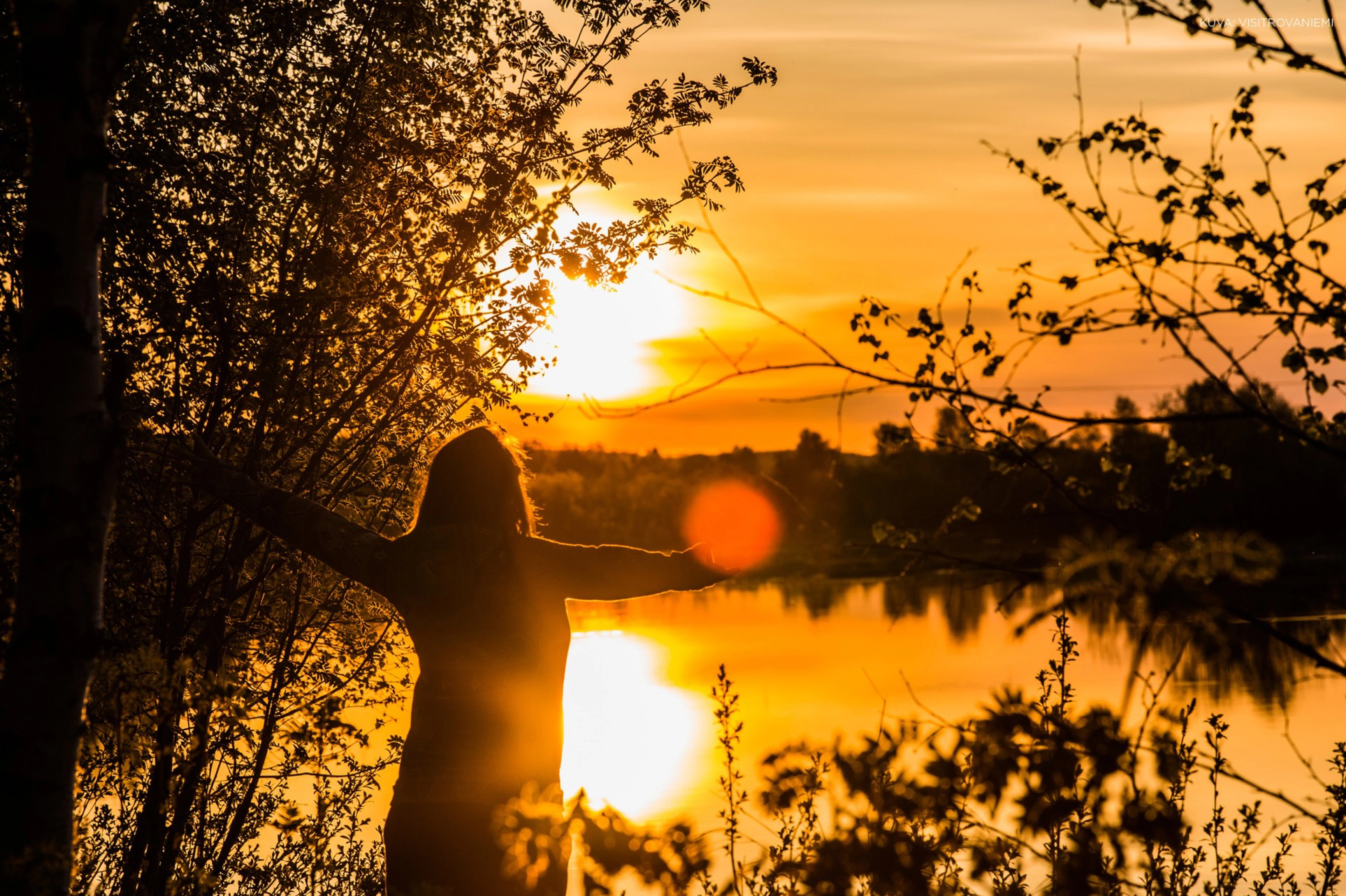 Yöttömän yön aurinko loistaa jokimaiseman yllä värjäten maiseman kultaiseksi, ihminen seisoo rannalla ottaen valoa avosylin vastaan