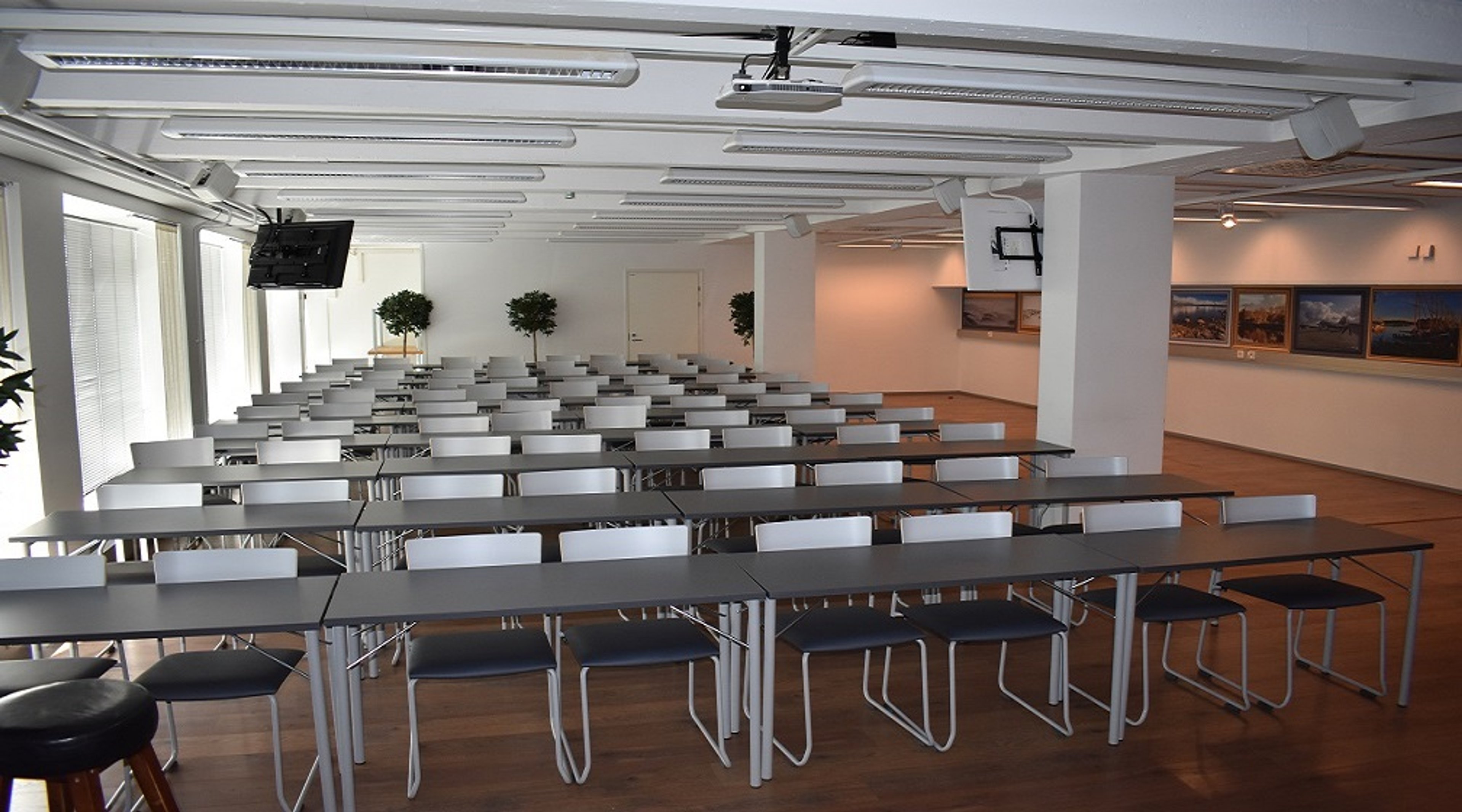 Bright meeting room with long tables and chairs arranged in rows. Wooden floor, small potted trees, and a display area with framed pictures on the wall. Ceiling projector installed.