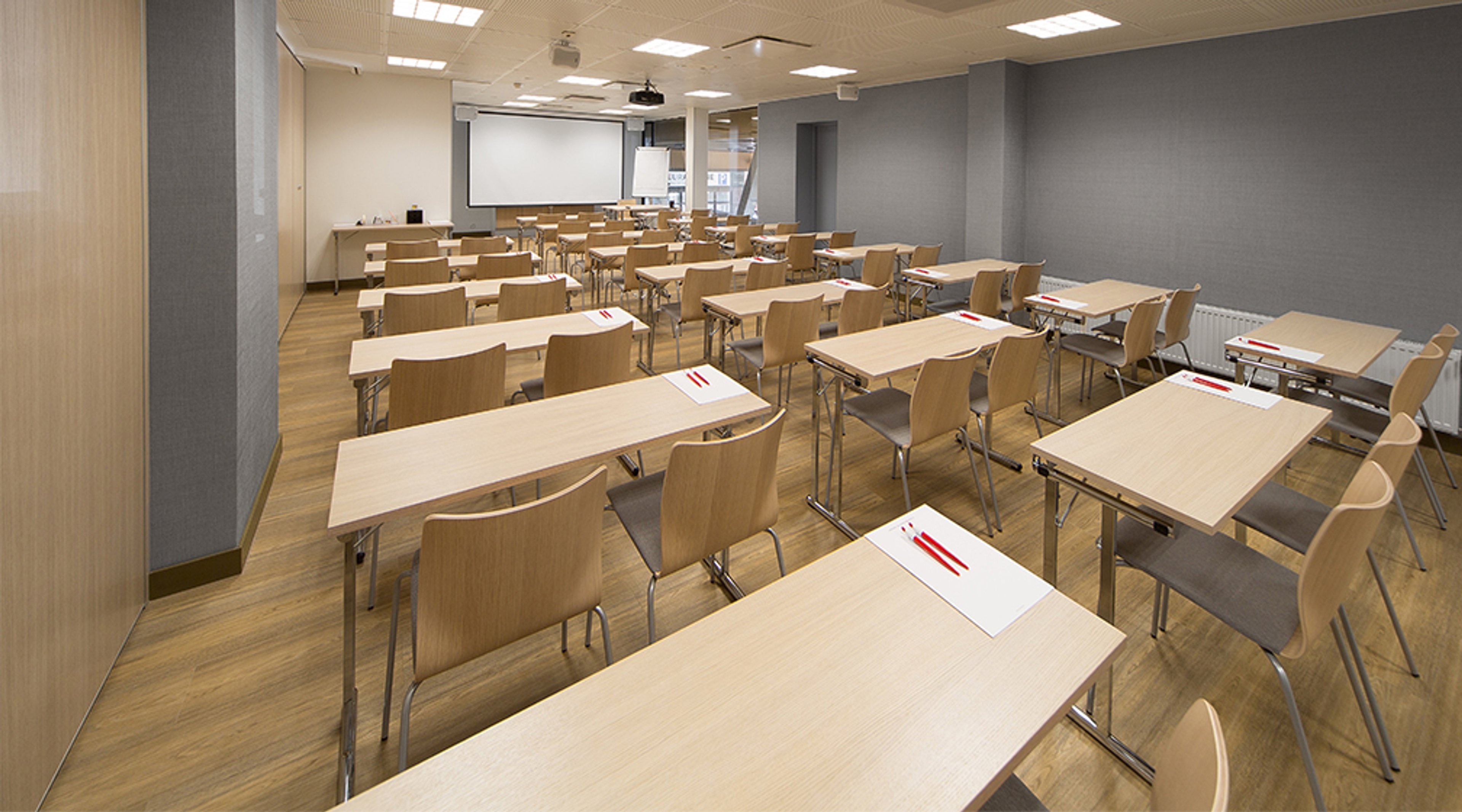 Modern classroom with wooden tables and chairs, projector and screen at the front, light wooden floor and even lighting.