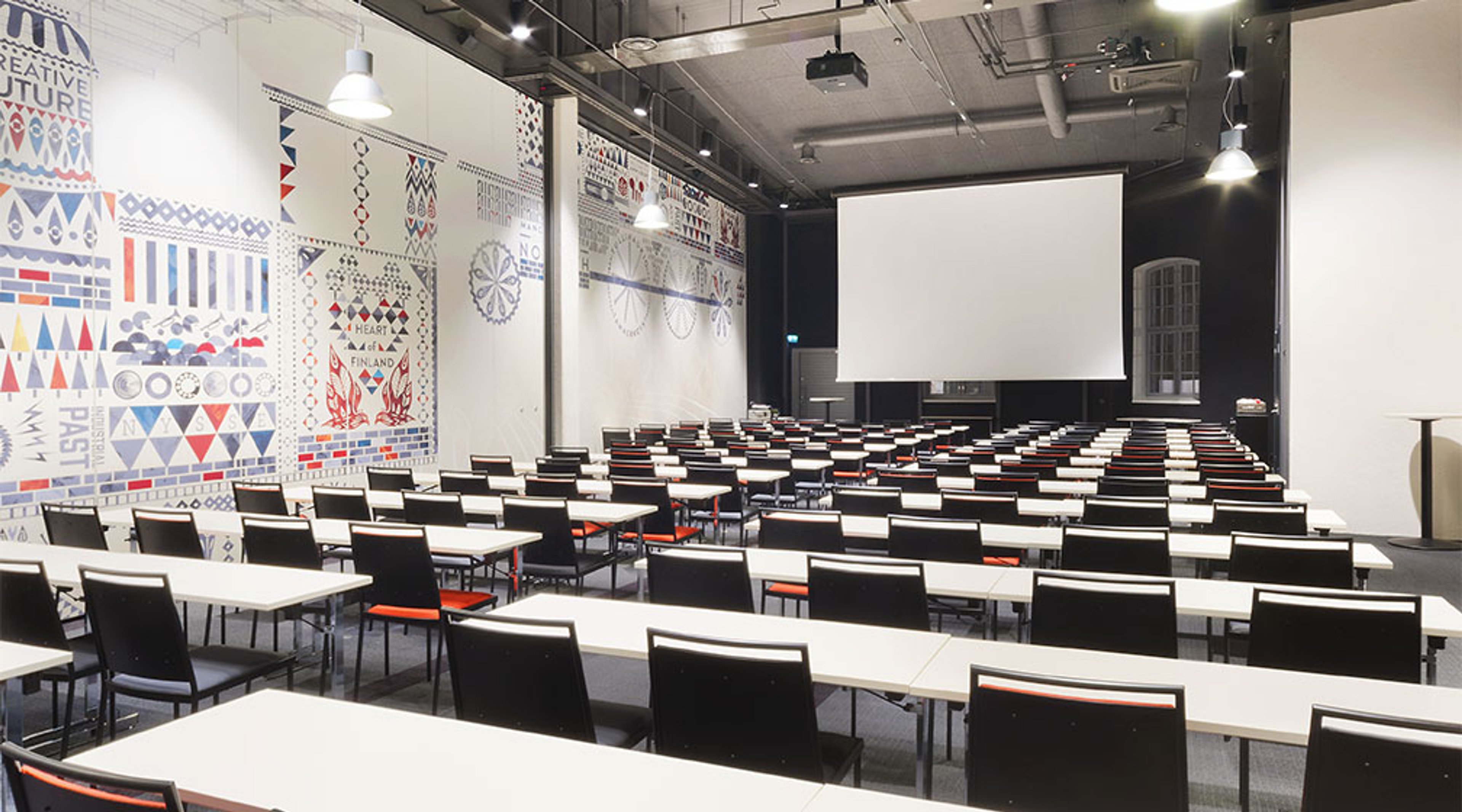 Moderni auditorio pöytäriveillä, joissa mustat tuolit ja valkoiset pöydät. Seinällä värikkäitä geometrisia kuvioita ja edessä valkoinen projektoriruutu.