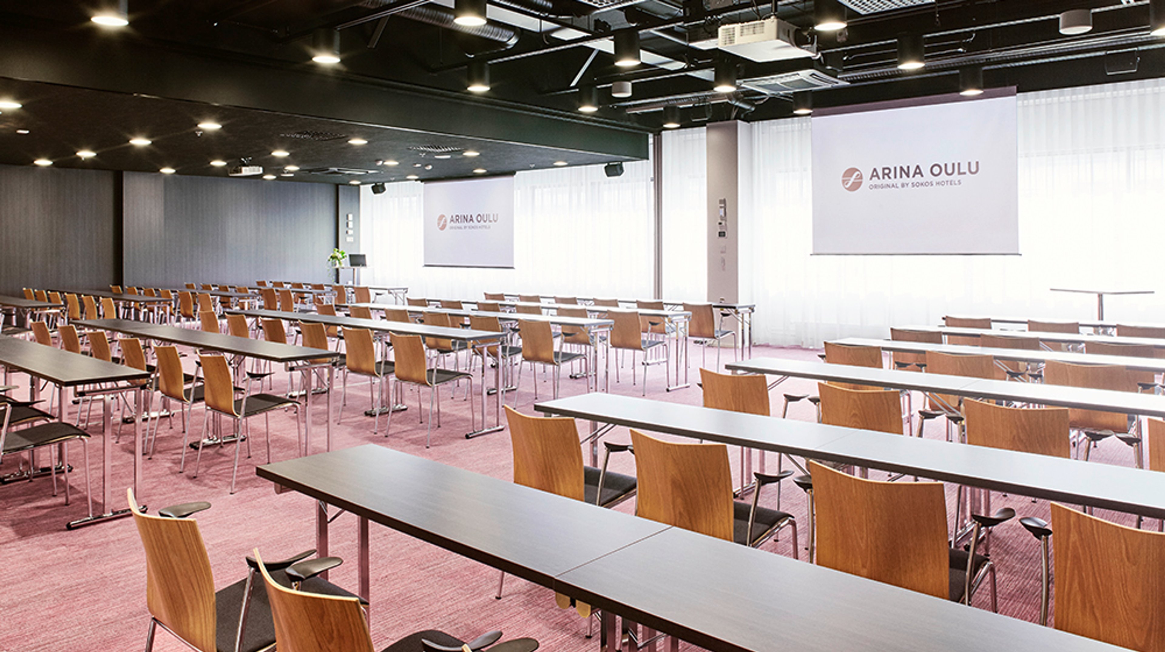 Spacious conference room with long tables and wooden chairs, pink carpeting, and two large projection screens.