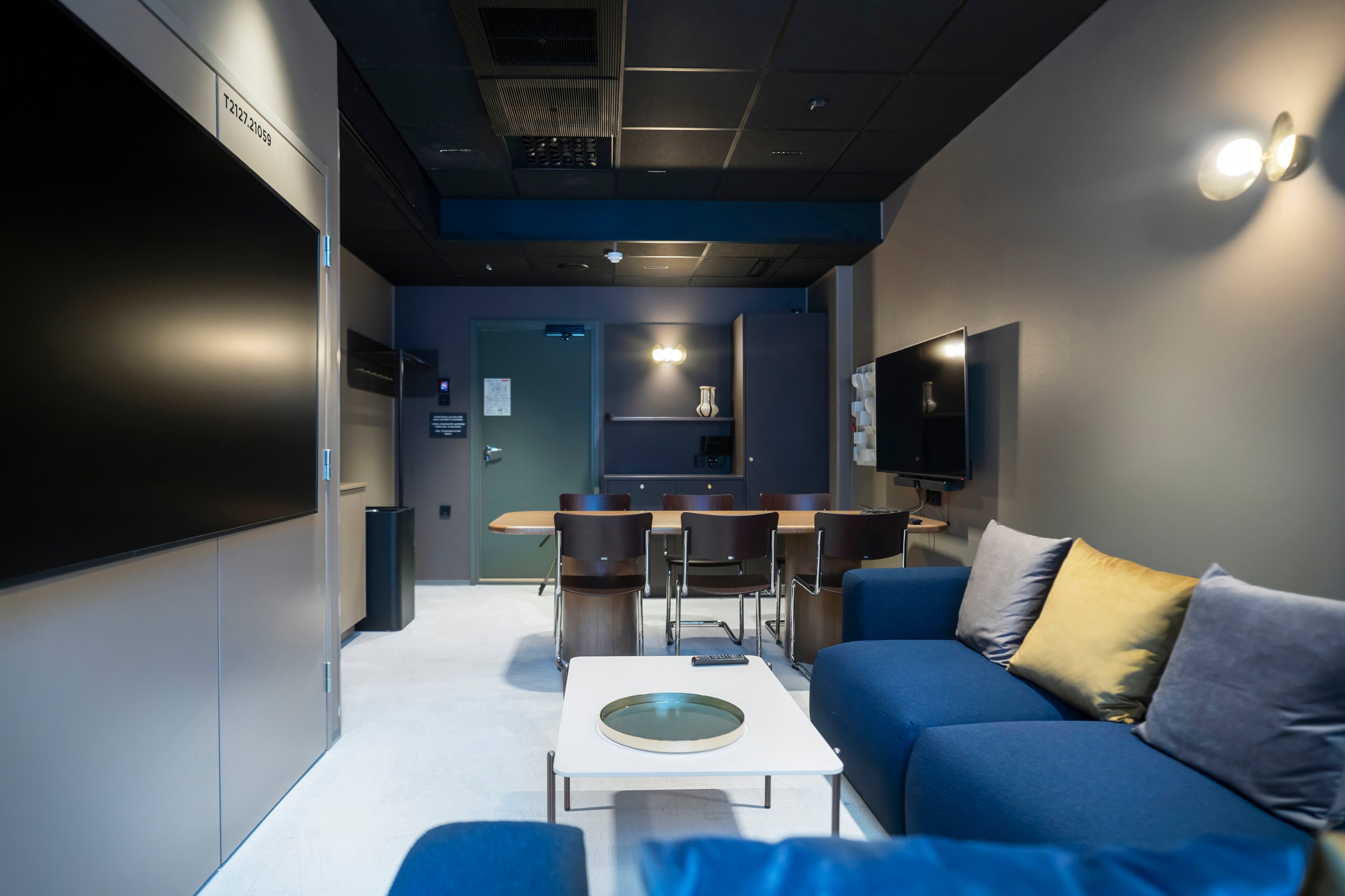 Modern meeting room with a blue sofa, white coffee table, chairs around a table, and two screens on the walls. Level floor and good lighting.