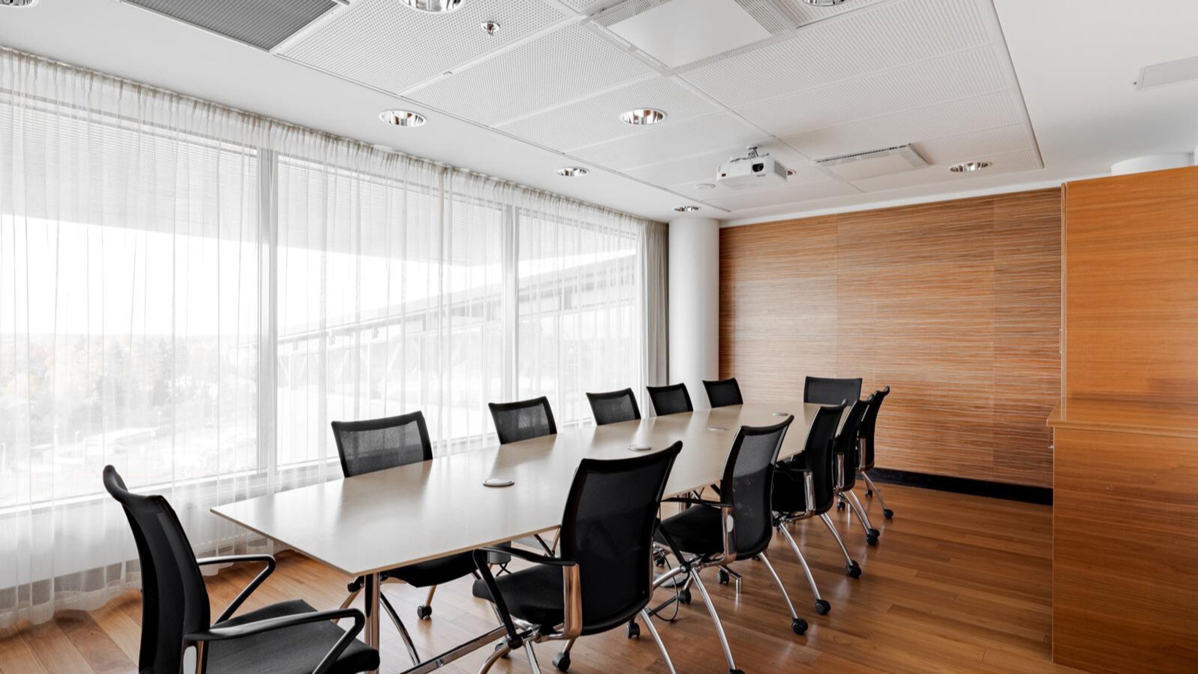 Modern conference room with long white table, black chairs, wooden flooring, wood-paneled wall and large windows with sheer curtains.