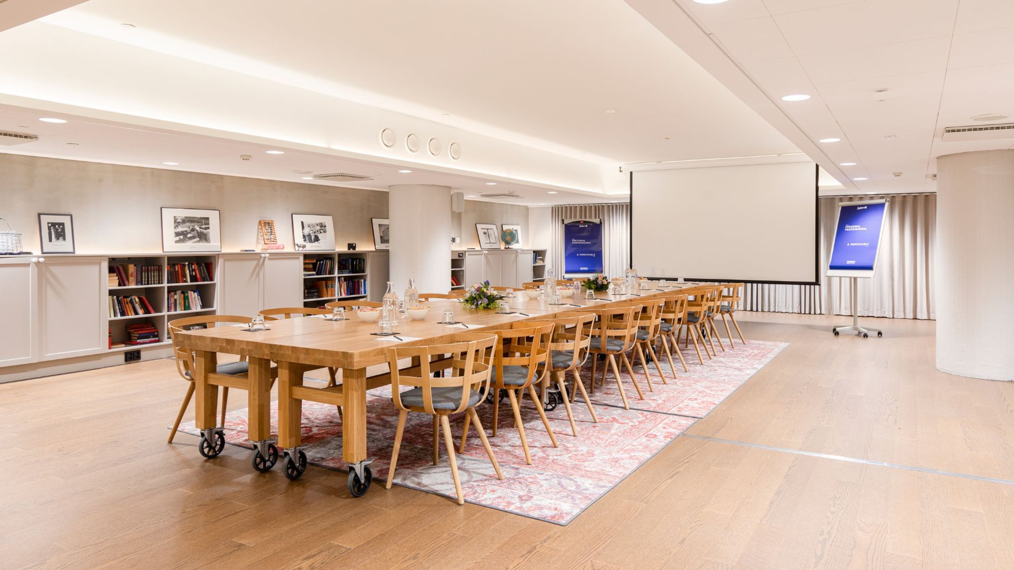 Kokoushuone, jossa on pitkä puinen pöytä pyörillä, tuolit, projektiokangas, näyttö ja kirjahyllyjä. Puulattia ja matto pöydän alla.