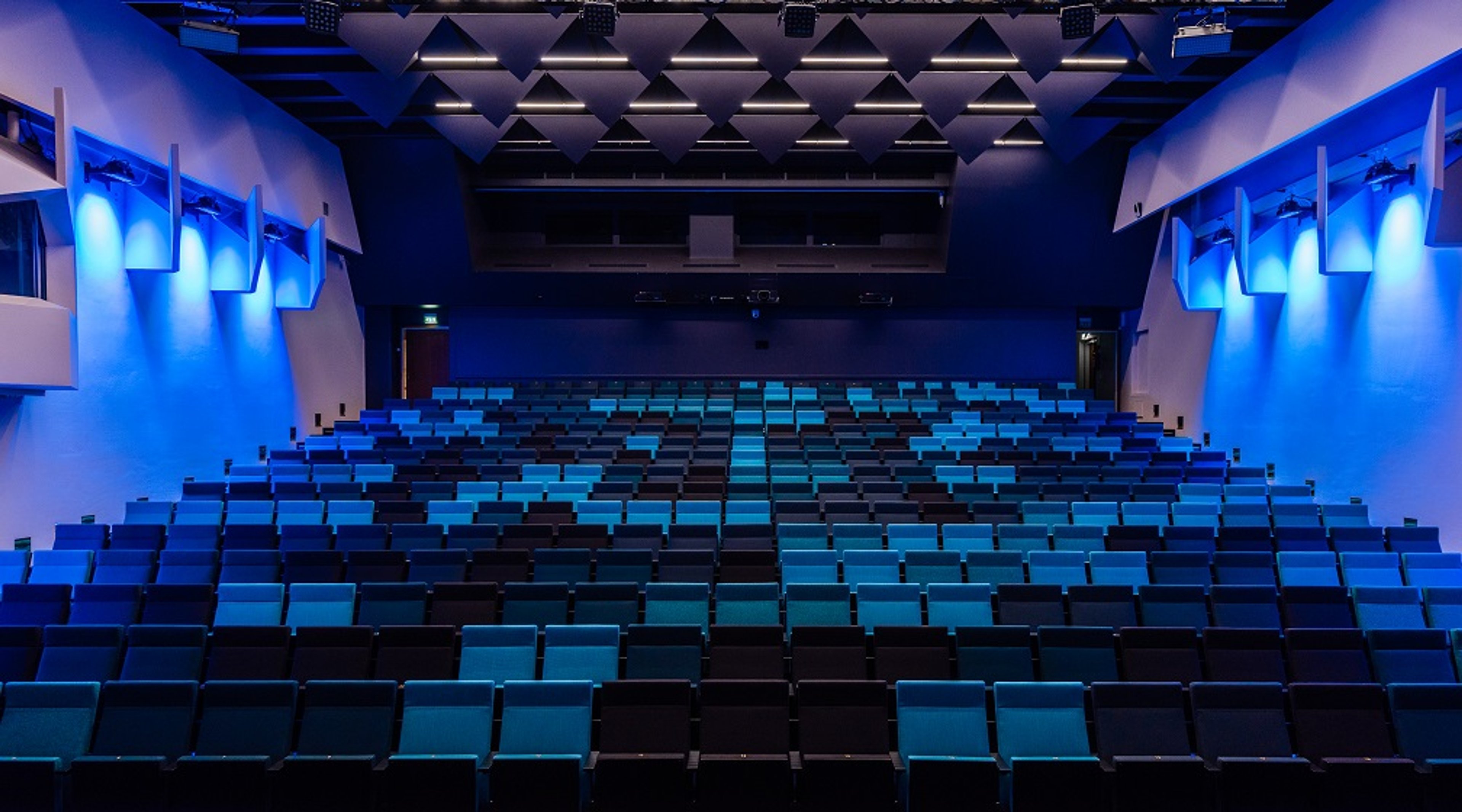 Modernt auditorium i blå belysning med stigande sittplatser och akustikpaneler i taket.