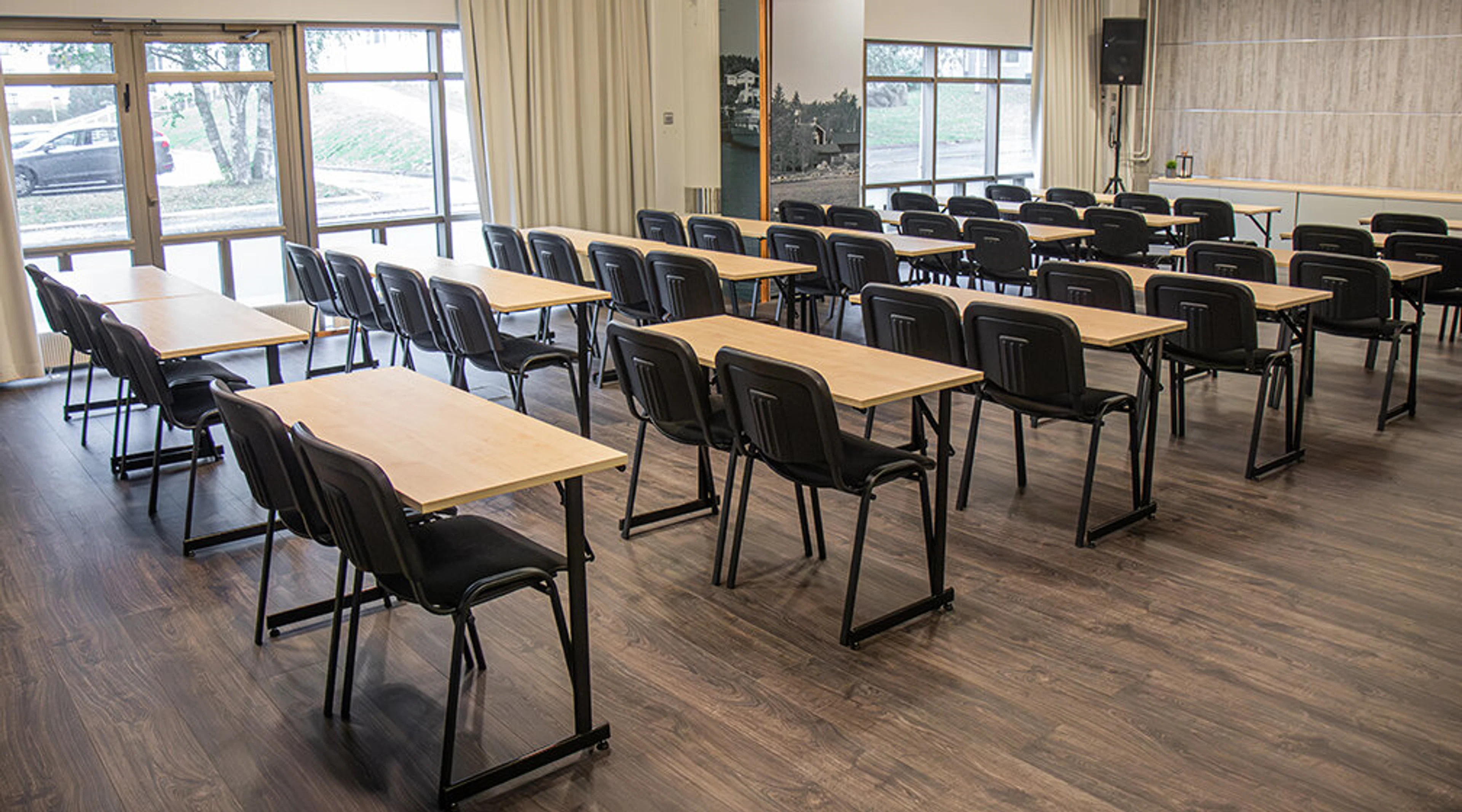 Meeting room with wooden floors, light-colored tables and black chairs arranged in rows. Glass walls allow natural light into the space.