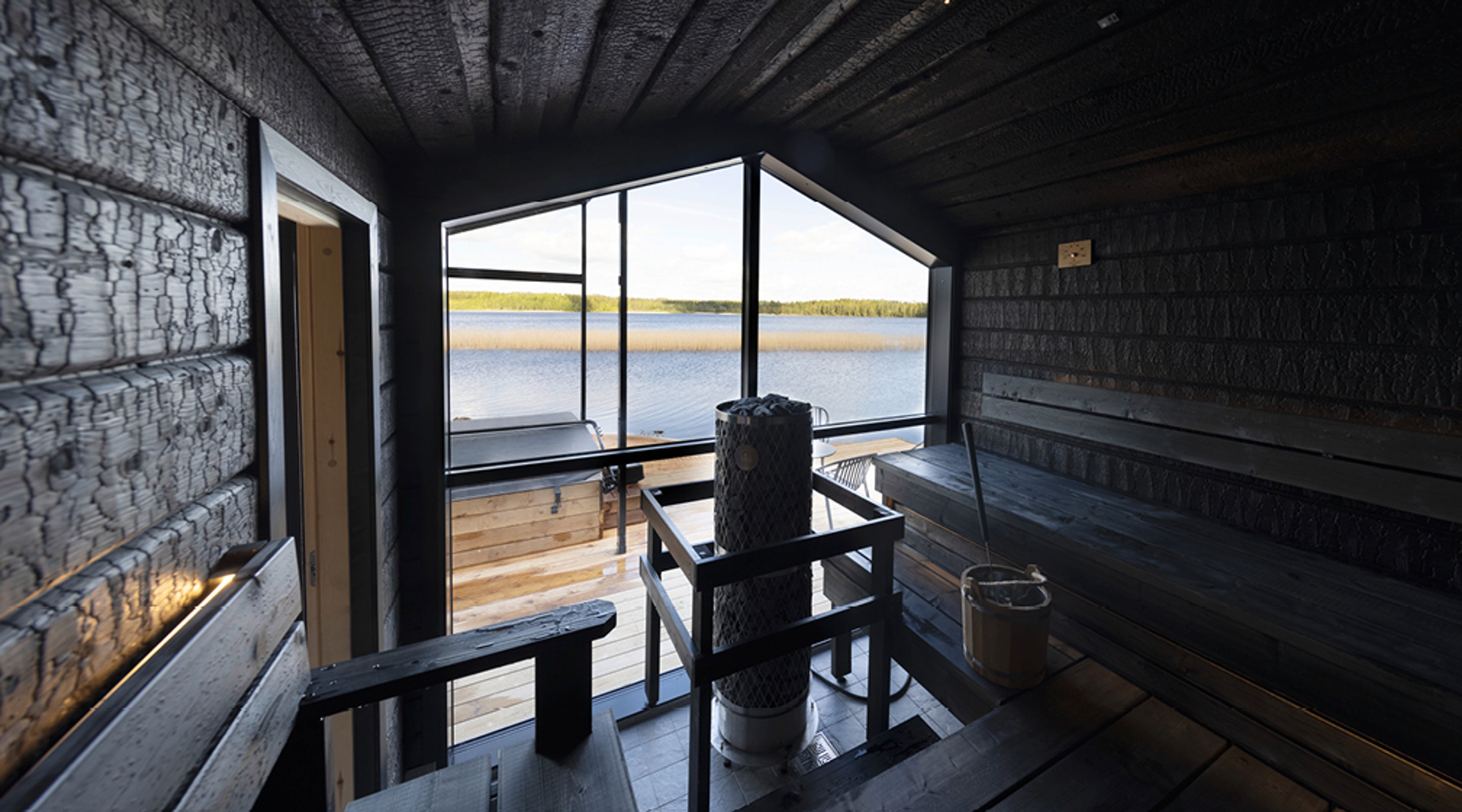 Moderni tumma sauna järvinäkymällä. Keskellä kiuas ja puiset lauteet. Suurista ikkunoista avautuu näkymä tyynelle järvelle ja metsämaisemaan.