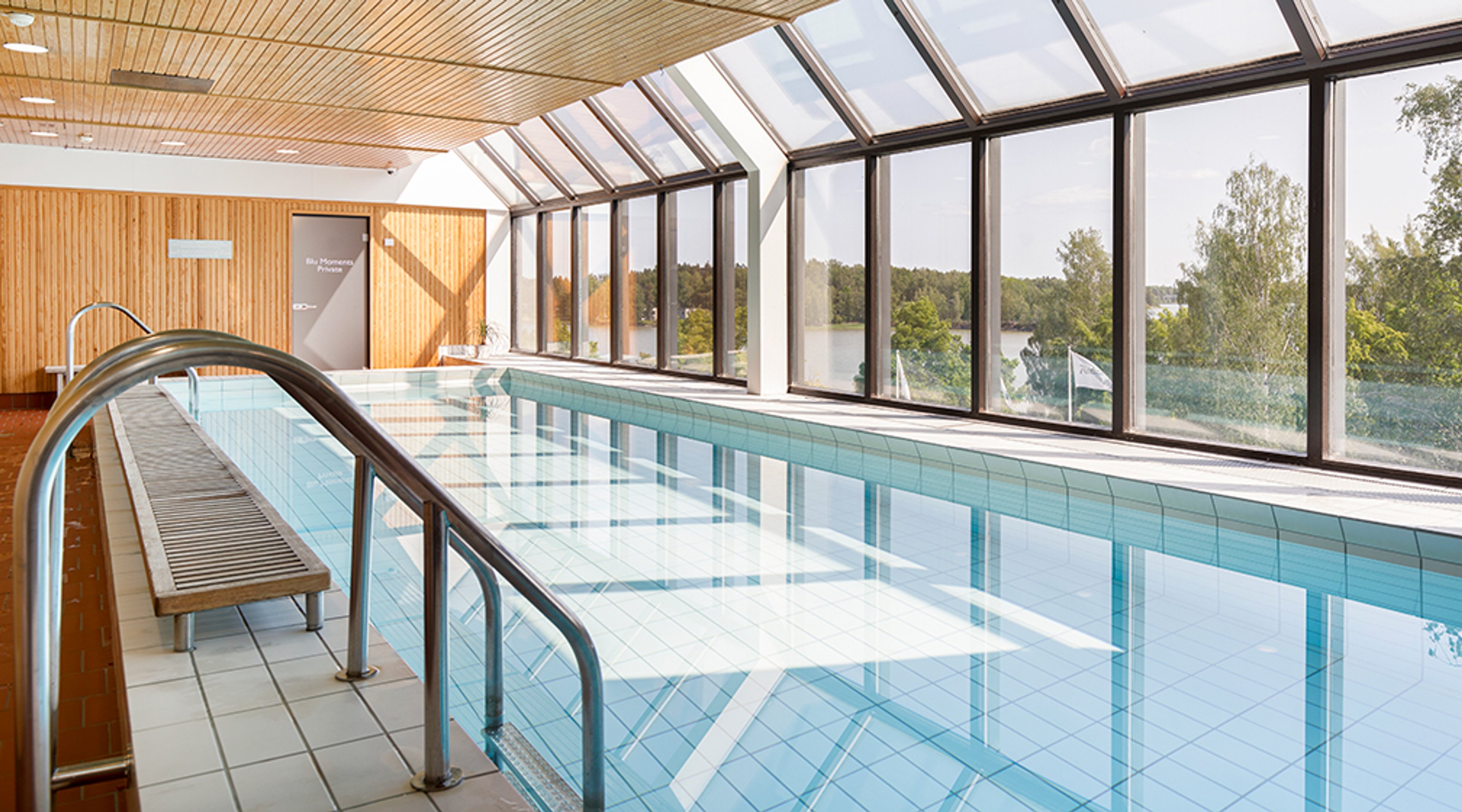 Inomhuspool med träpaneltak och stora fönster som visar sjöutsikt. Metallräcke och bänk vid poolkanten.