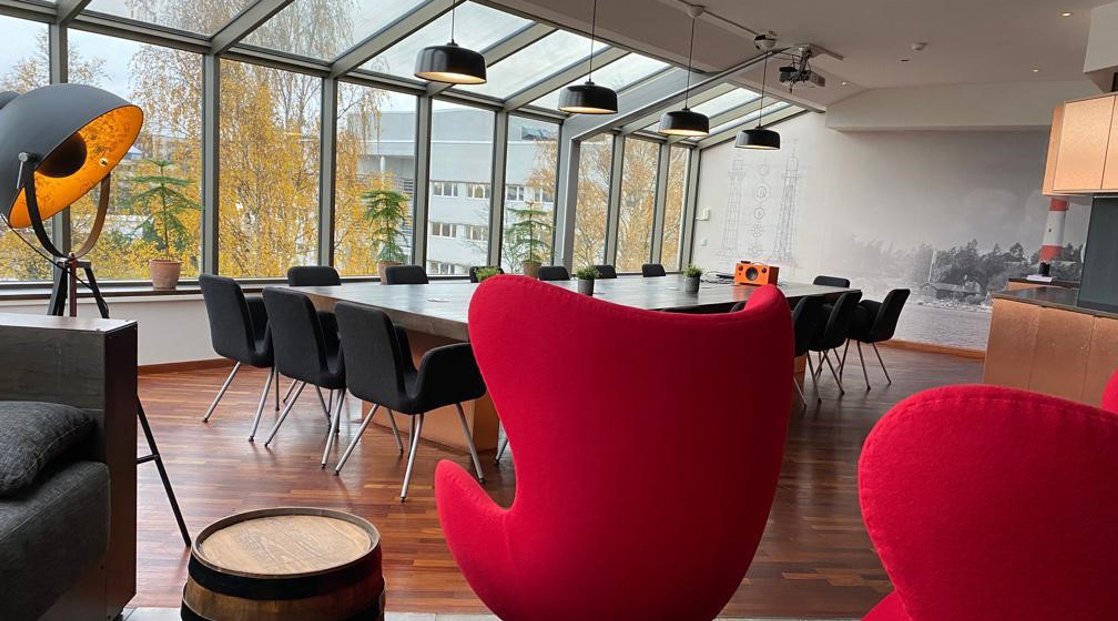 Modern meeting room under glass ceiling with long table, black chairs and red armchairs. Wooden floor and view of autumn trees.
