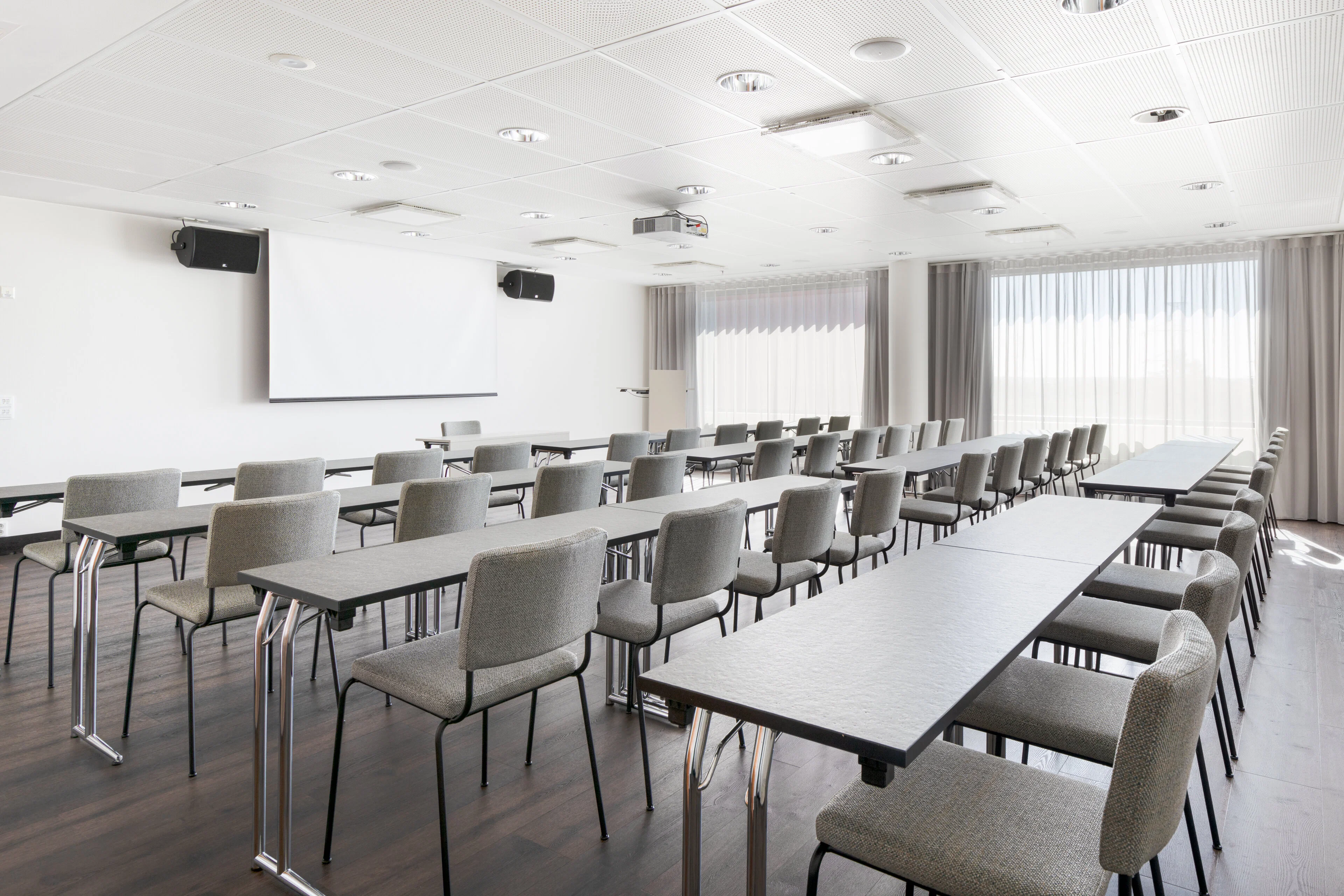 Bright conference room with long rows of tables, grey chairs, white projection screen and grey curtains covering windows.