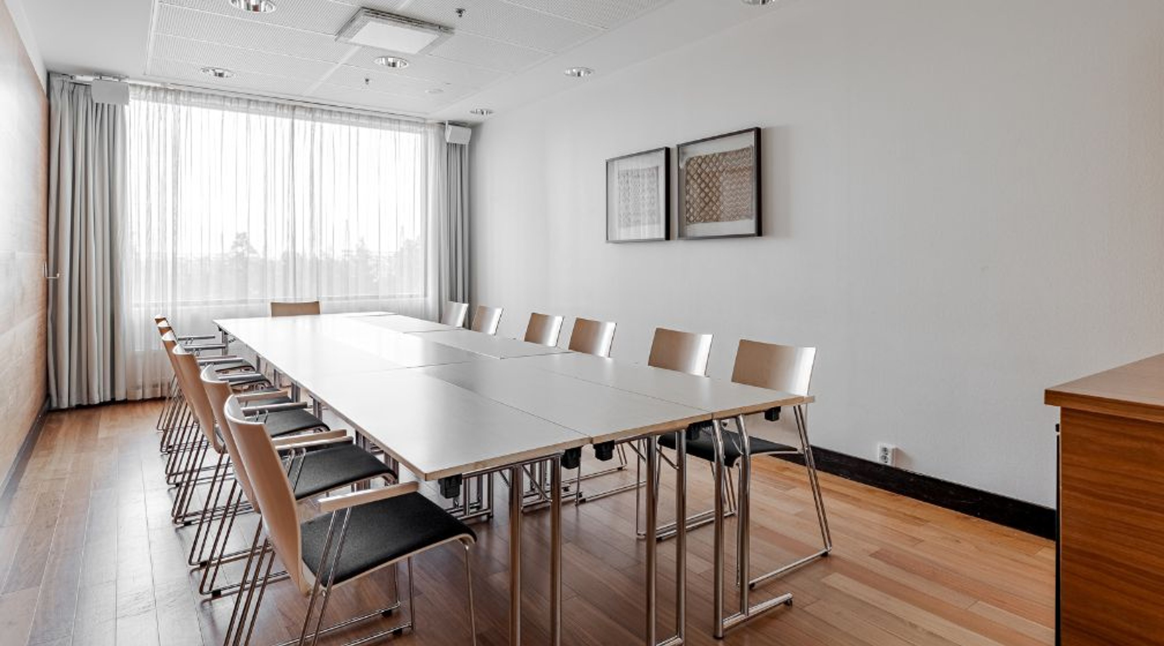 Meeting room with long white table, light-colored chairs and large curtains. Wooden floor and two framed artworks on the wall.