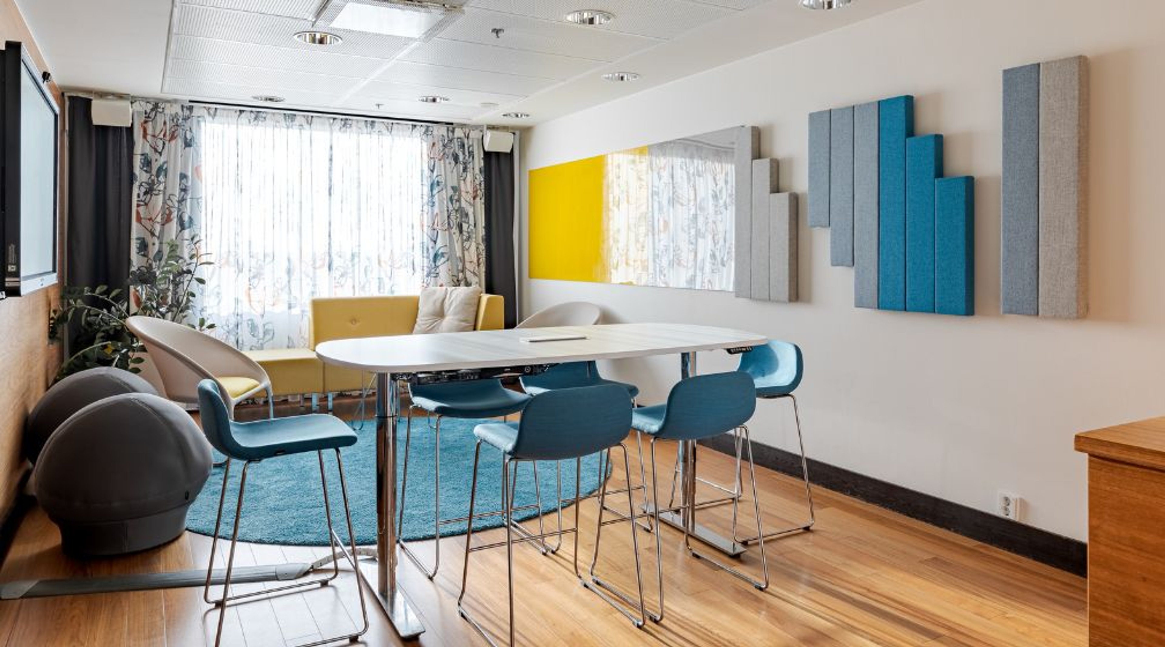 Modern meeting room with white table, blue bar stools, yellow sofa and acoustic panels on the wall. Wooden floor and large windows.