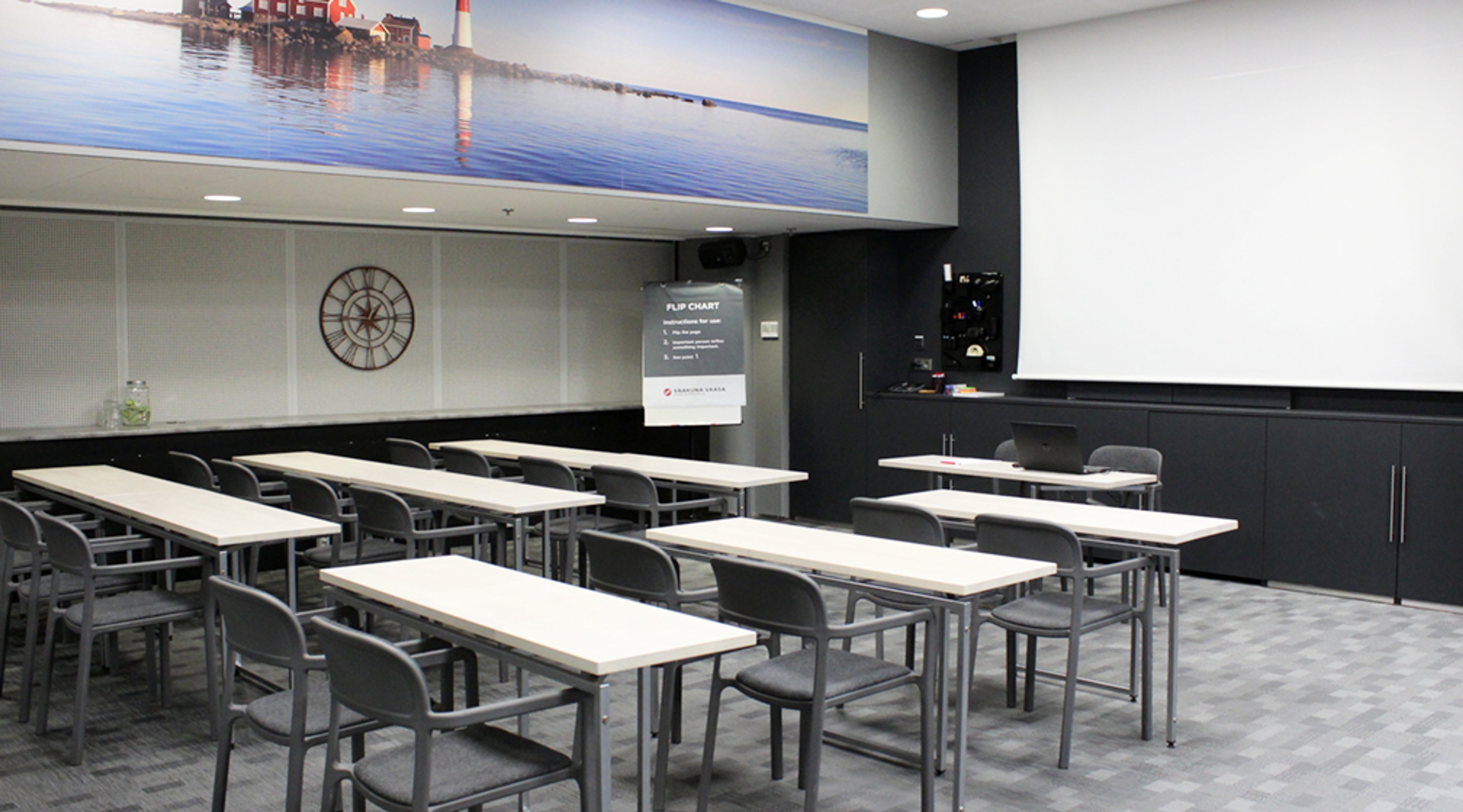Meeting room with white tables and gray chairs arranged in rows. A landscape image of waterfront and a clock on the wall.