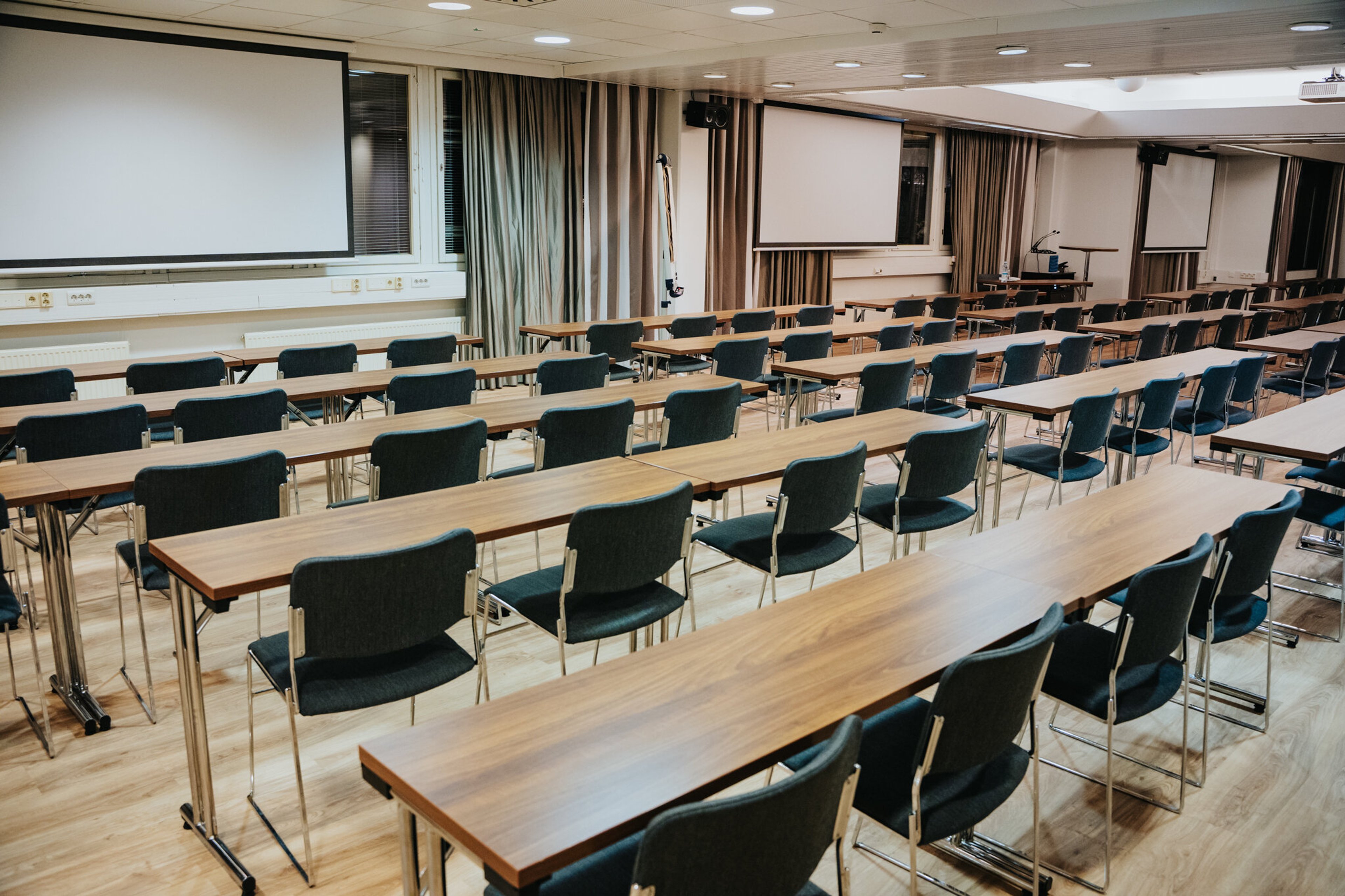 Auditorio, jossa pitkiä pöytiä ja tuoleja riveittäin. Edessä valkokankaita ja taka-alalla verhoja. Tasainen puulattia ja hyvä valaistus.