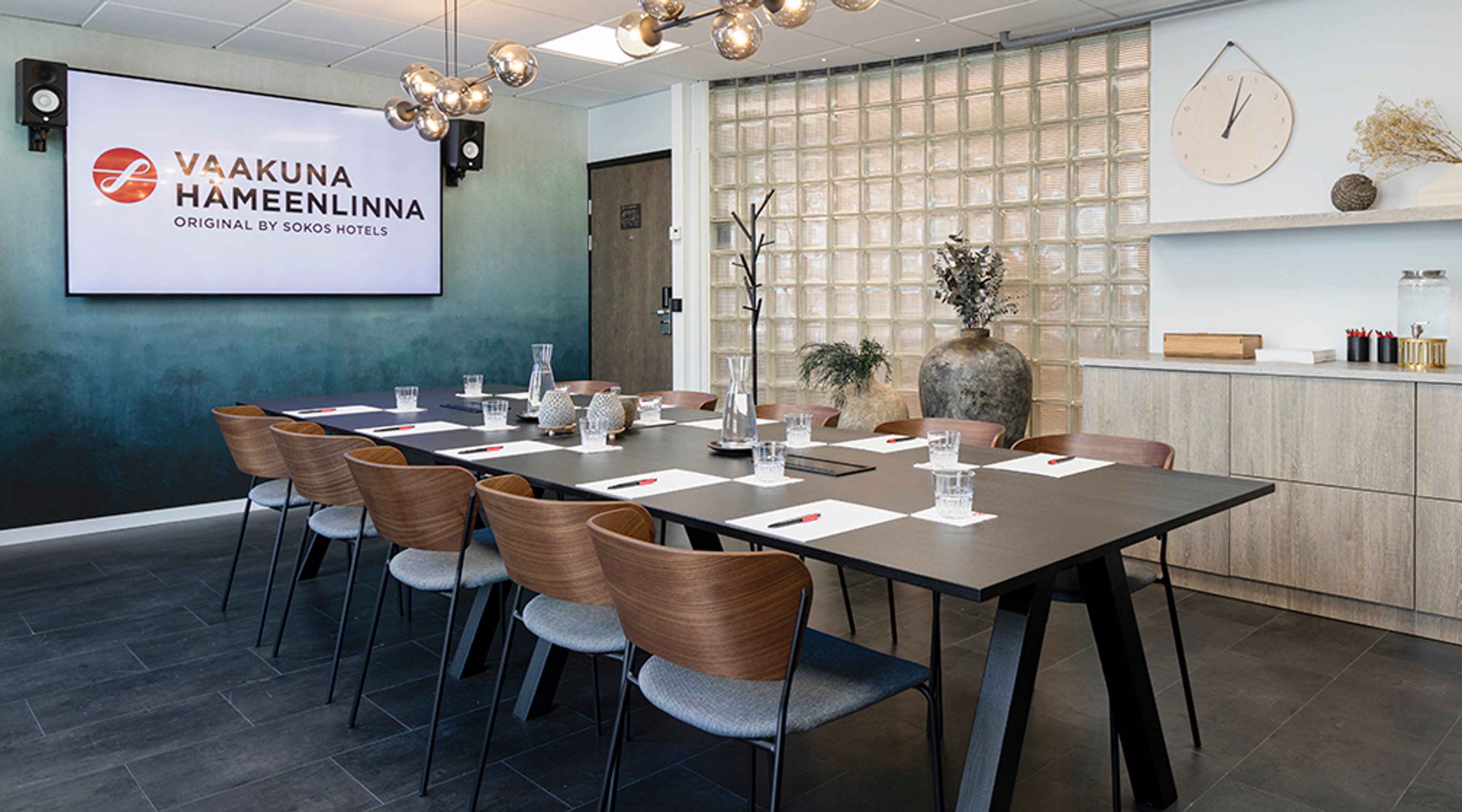 Bright meeting room with long table and wooden chairs, screen on wall. Modern interior with glass block partition and dark tile flooring.