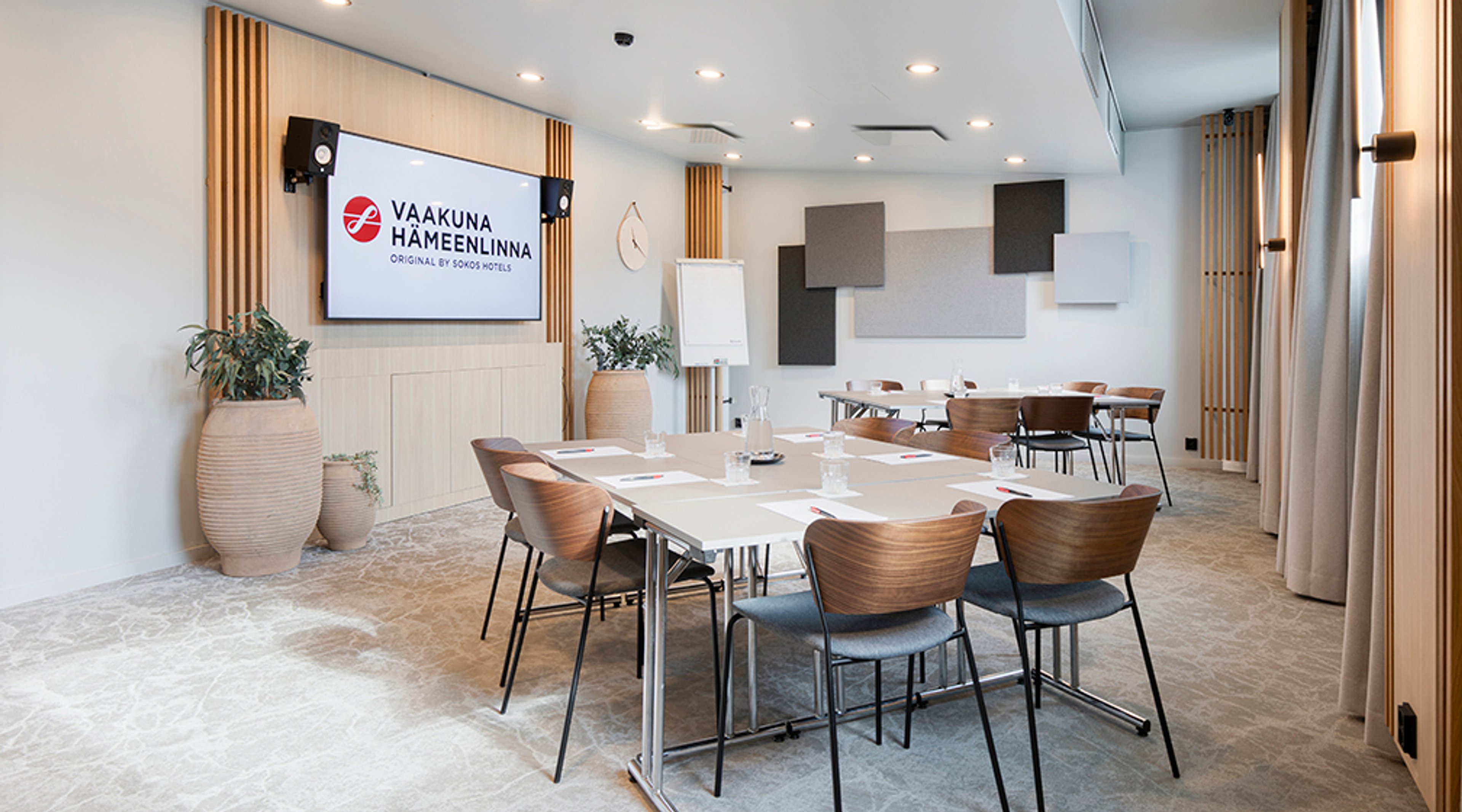 Modern meeting room with light walls, wooden details and stylish chairs. The space features a large display screen, flipchart and acoustic panels.