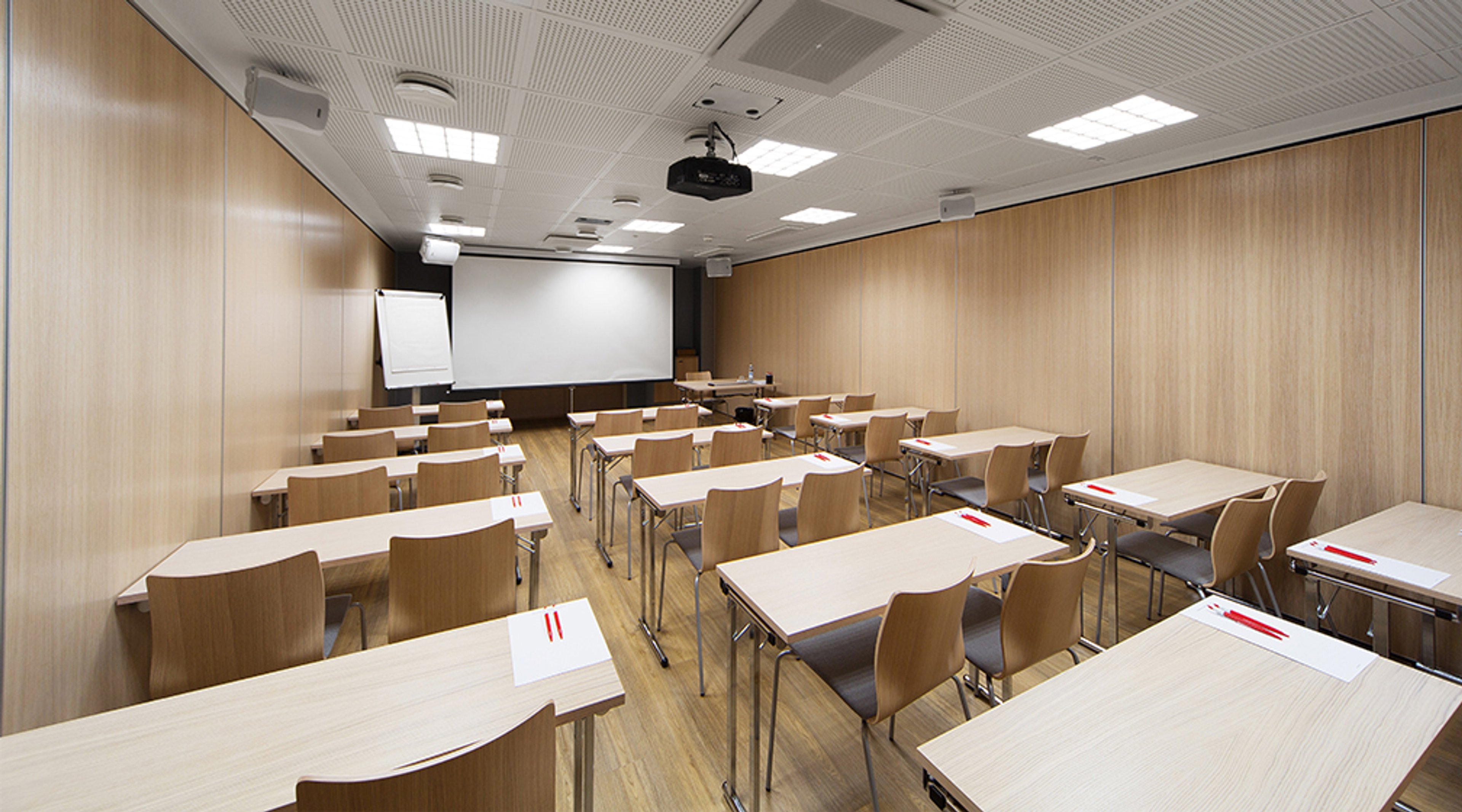 Meeting room with wooden walls, tables and chairs arranged in rows, projection screen ready for presentation, wooden floor and even lighting.