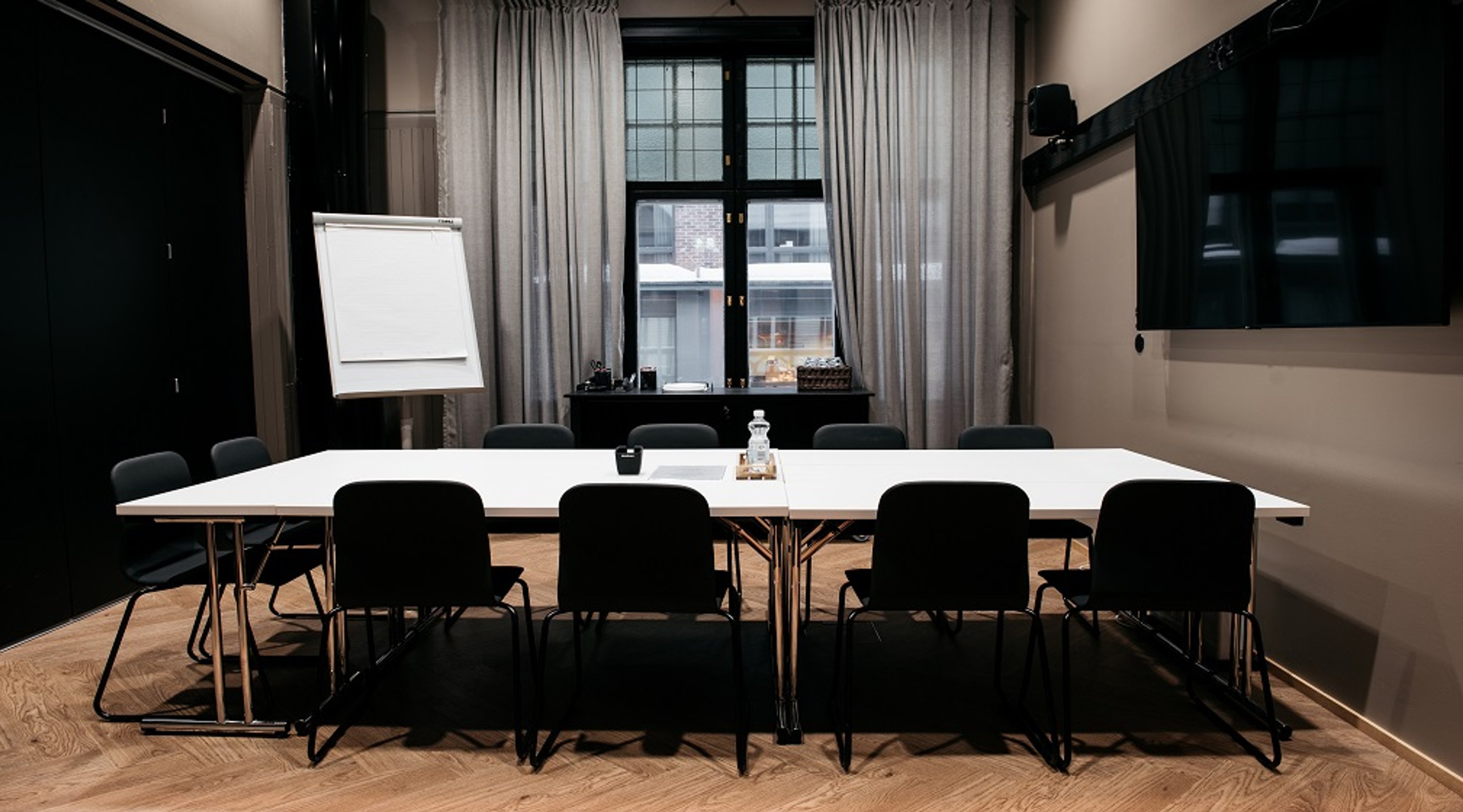 Modern meeting room with white table, black chairs, flipchart and TV. Wooden parquet flooring and light curtains at the window.