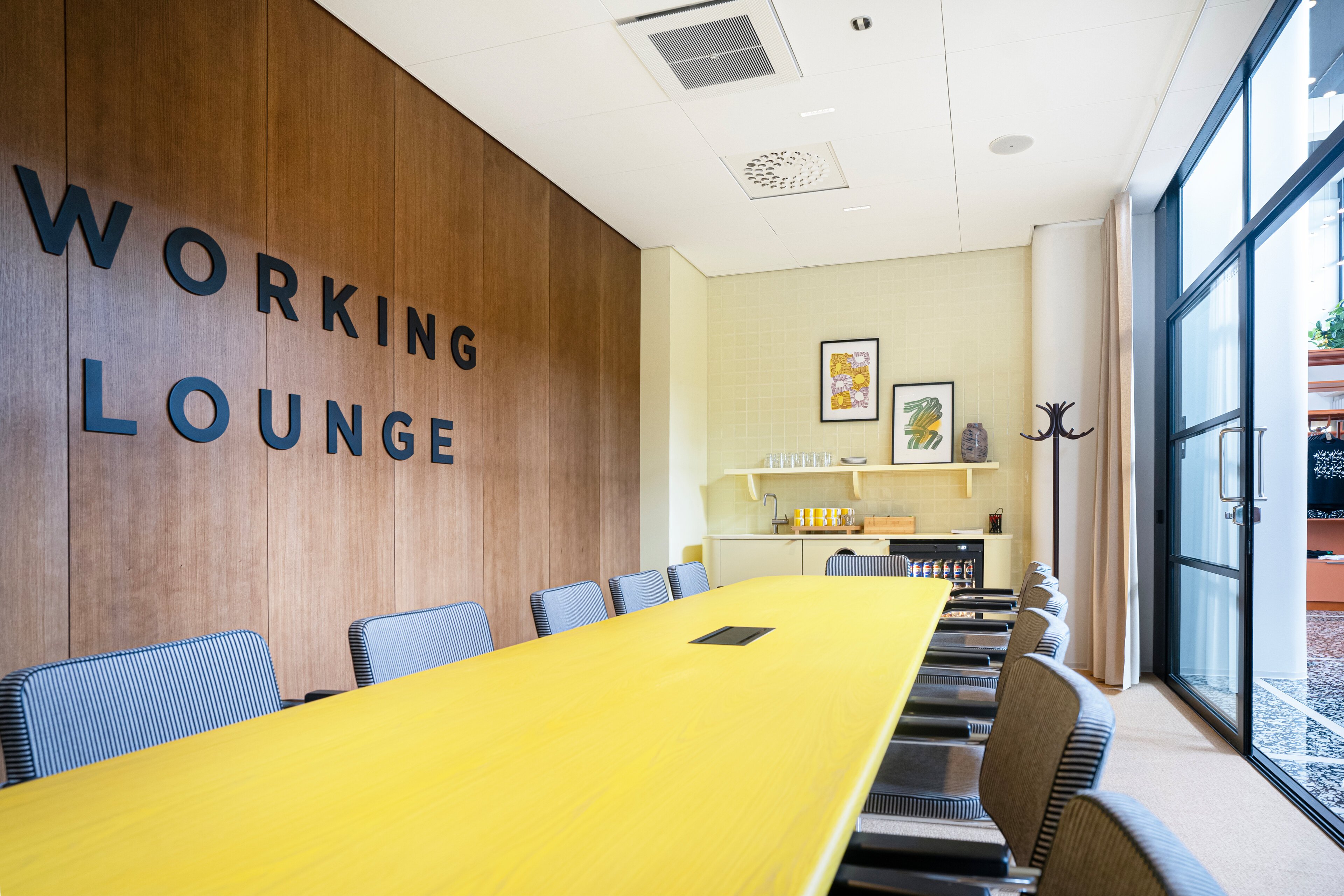 Modern collaborative space with yellow conference table, striped chairs, wooden panel wall, kitchenette area, and large windows. Level flooring and wide clearance areas.