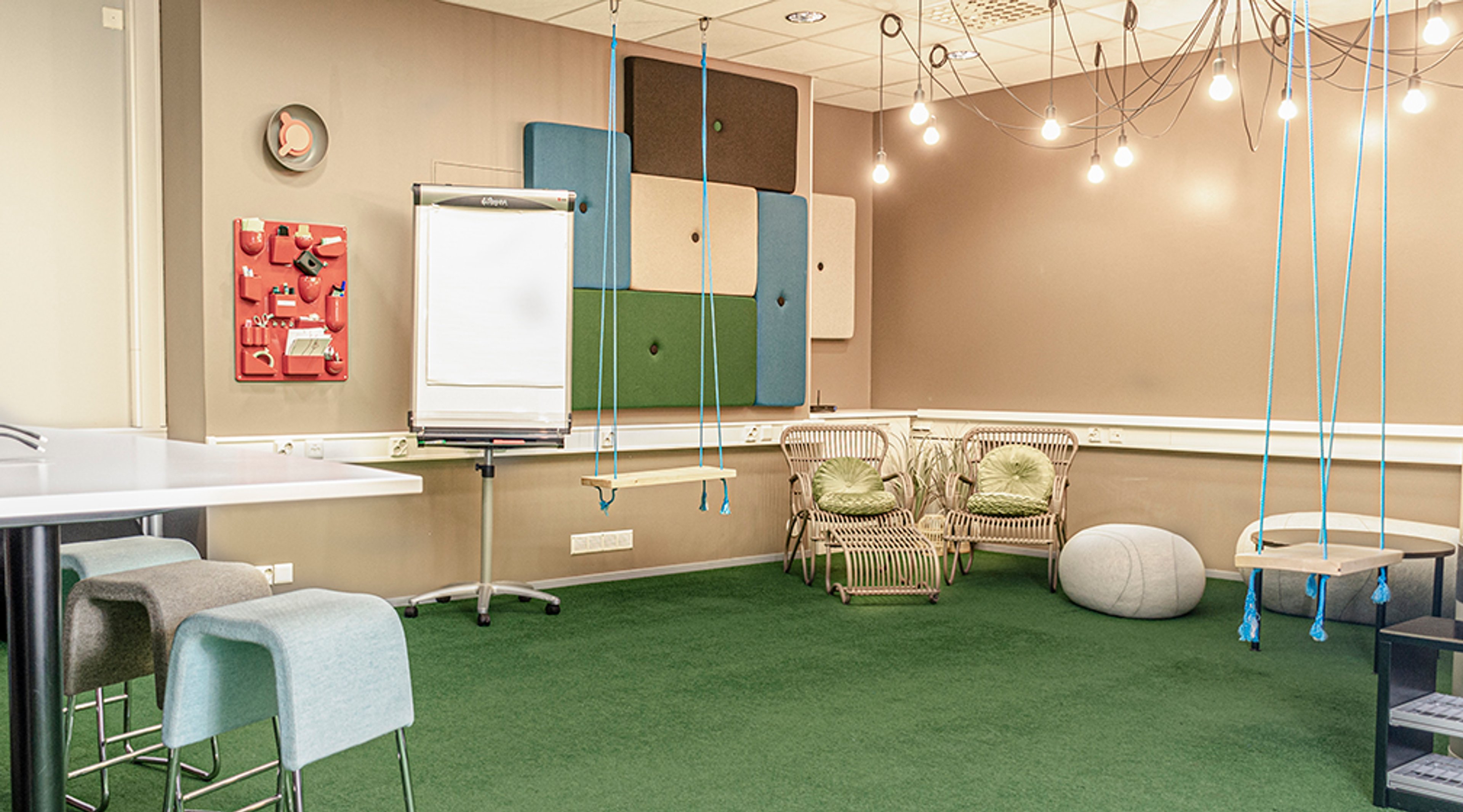 Creative meeting room with green carpet, hanging lights and swings. Features a flipchart, colorful acoustic panels, and rattan chairs.
