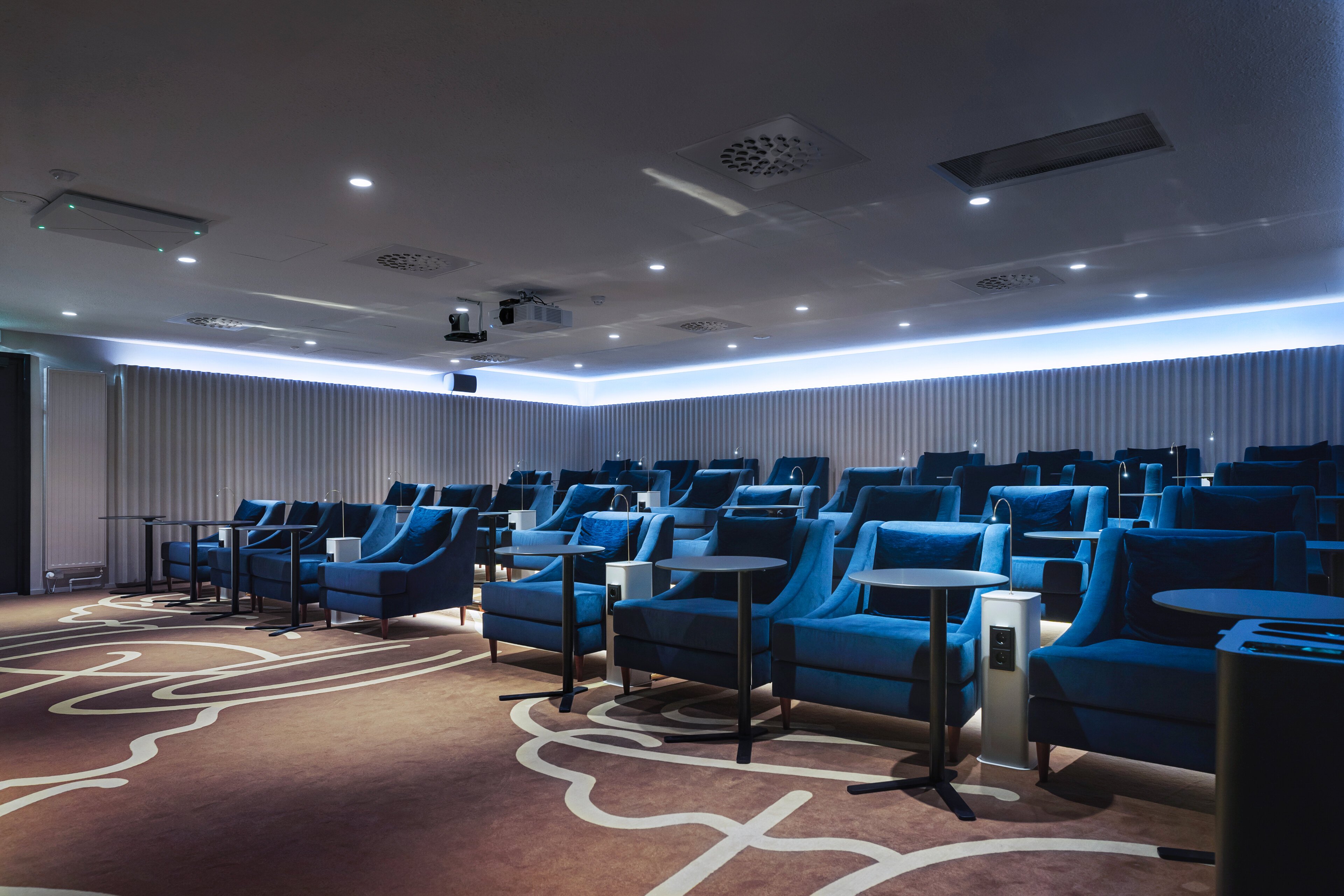 Modern auditorium with comfortable blue armchairs arranged in rows, small tables between seats, and decorative patterned carpet. Soft blue ambient lighting throughout.
