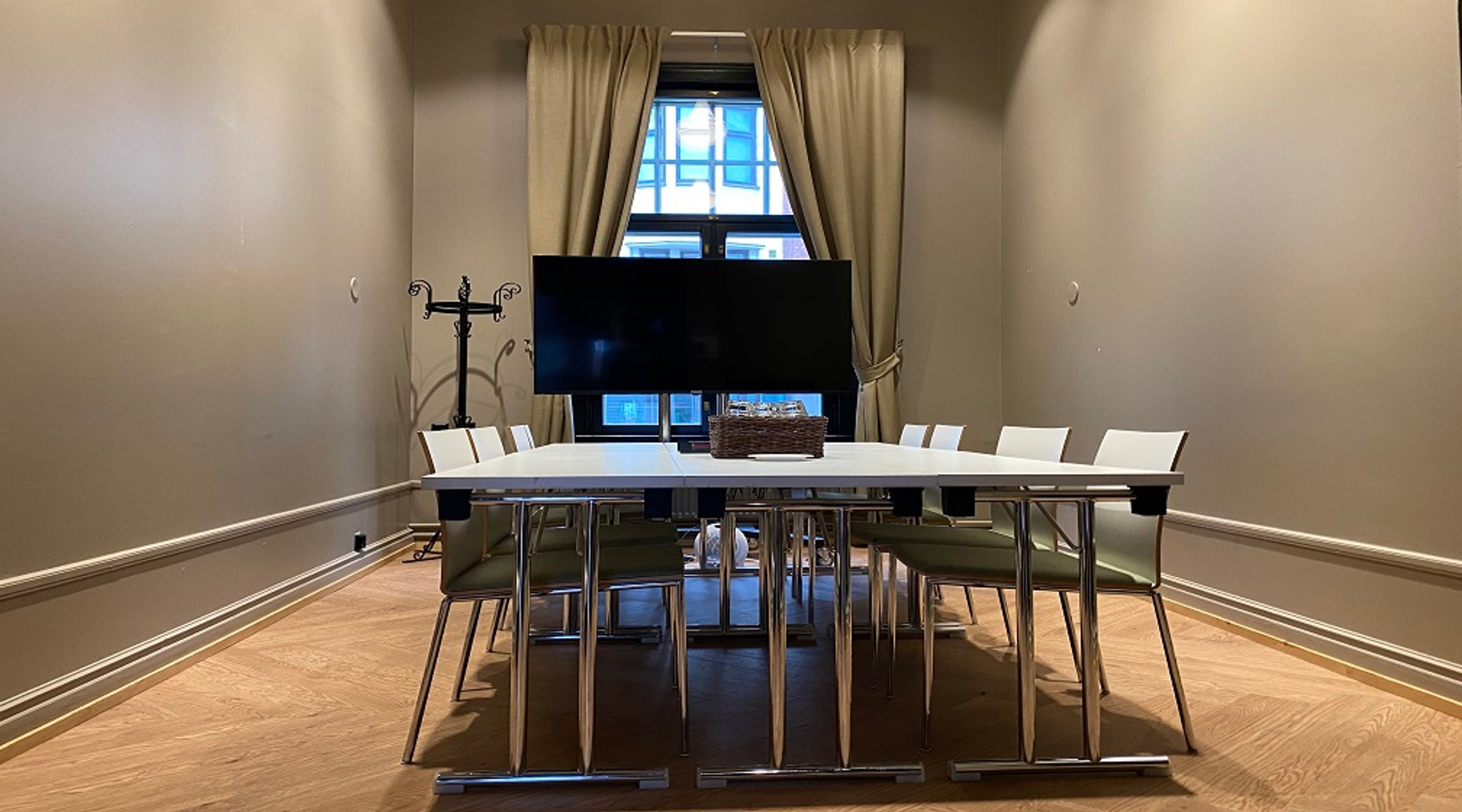 Meeting room with long white table and eight chairs. Wooden floor and a TV in front of the window next to beige curtains.