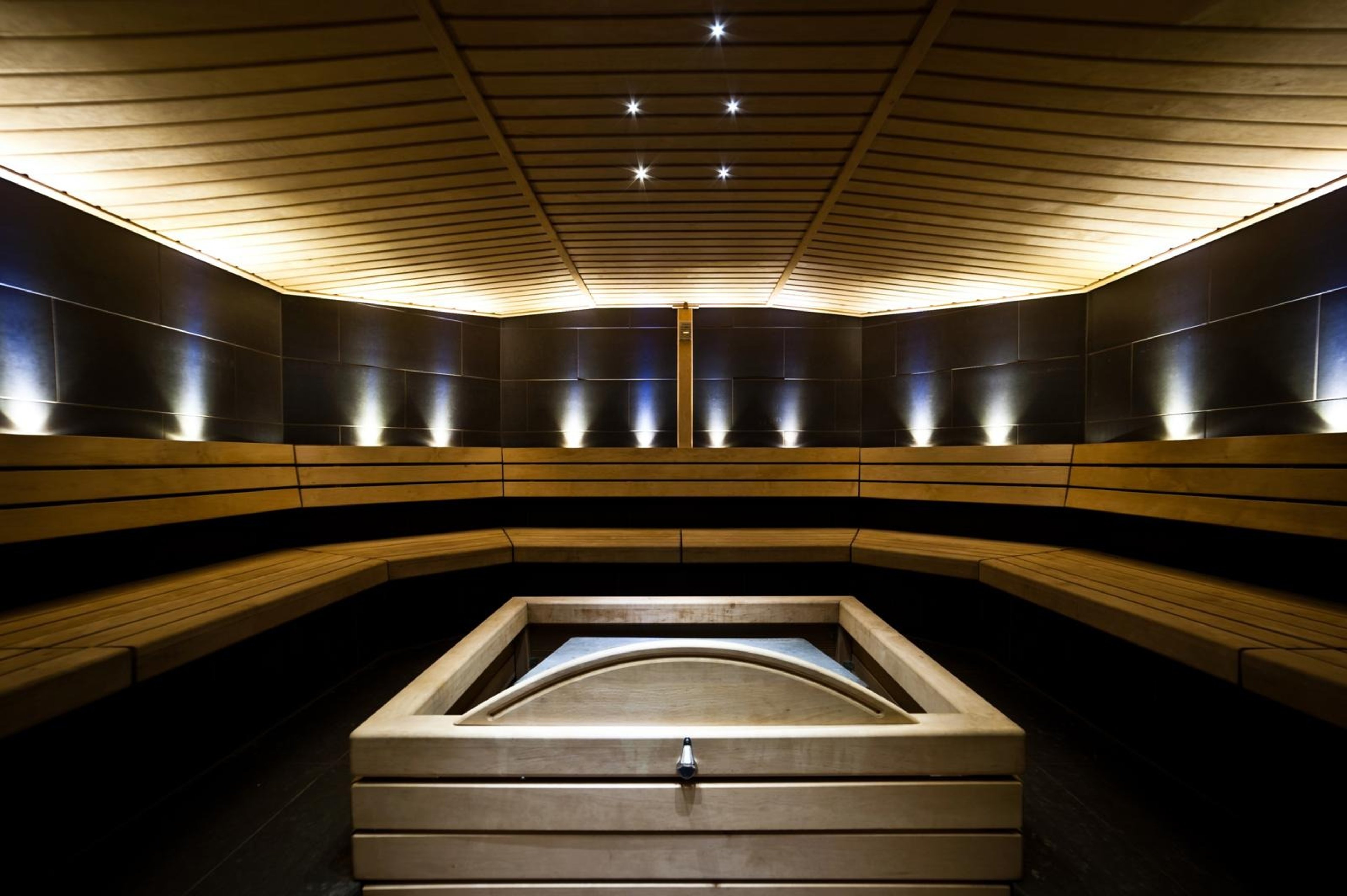 Moderni sauna, jossa on puiset lauteet, tummat seinät ja kattovalaistus. Keskellä on neliönmuotoinen kiuas.