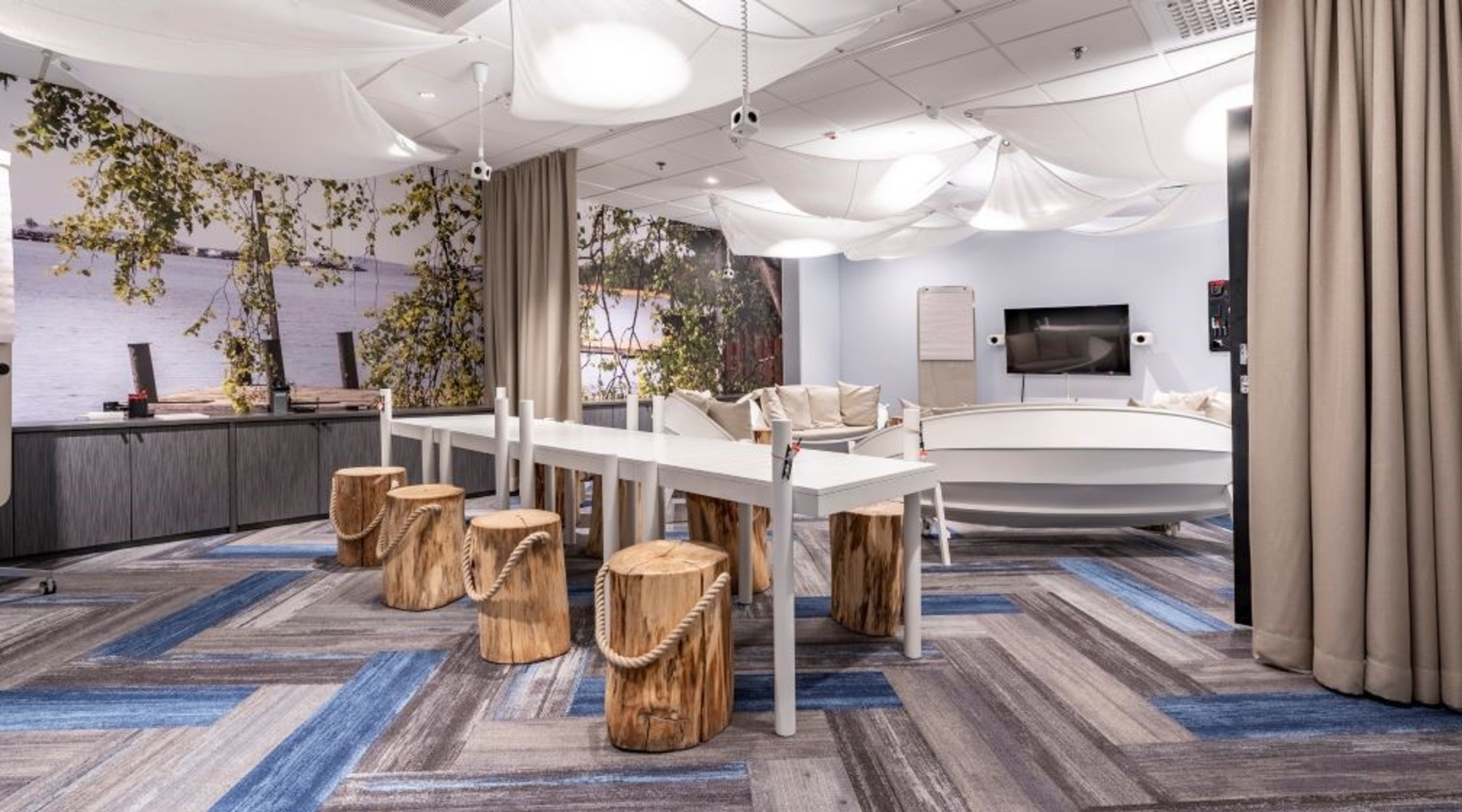 Modern meeting space with white table and wooden log seats, blue-toned carpet, nature imagery on walls and light-colored seating area in background.