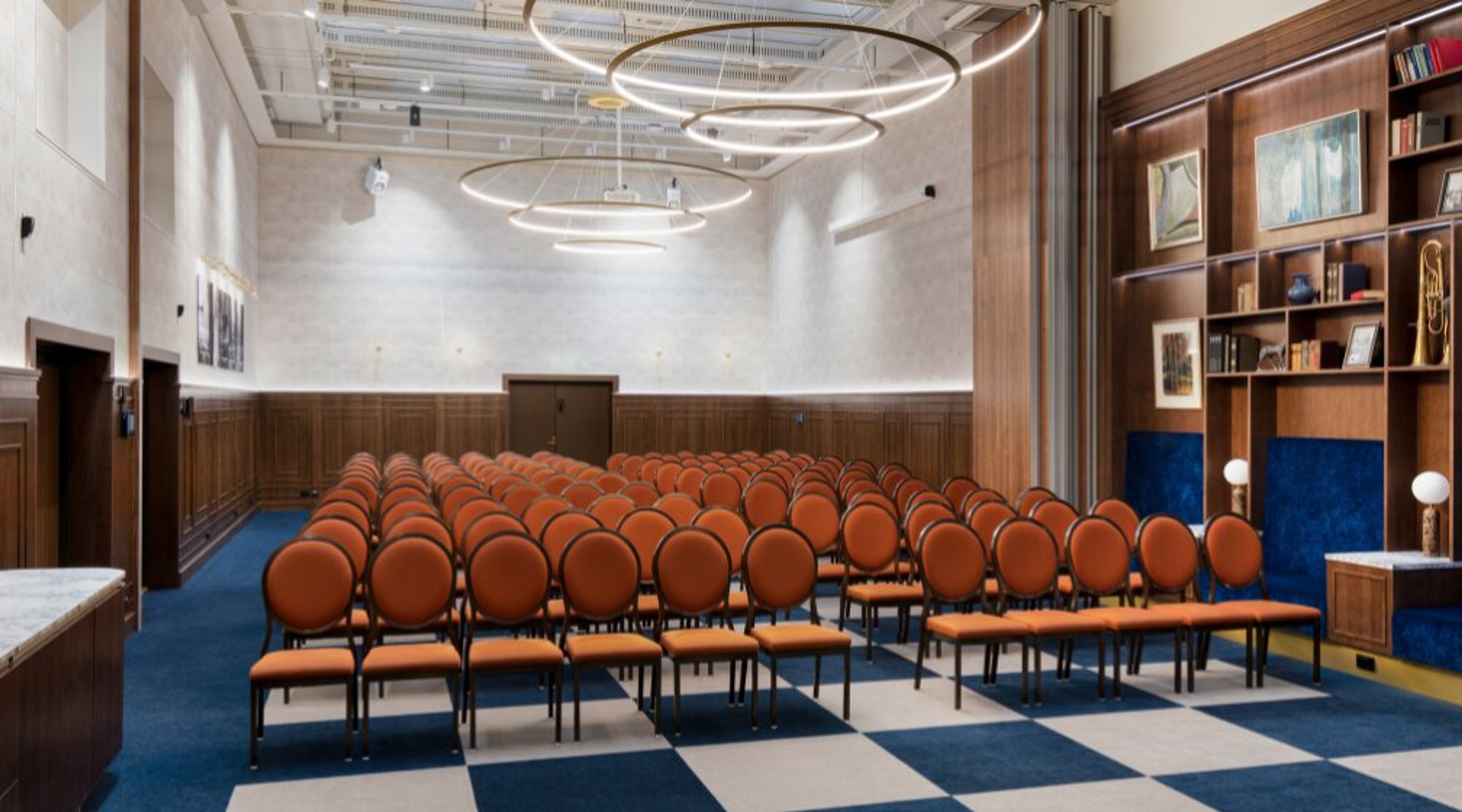 Modern auditorium with rows of orange chairs, blue carpet, wooden wall panels and circular ring lighting fixtures on the ceiling.