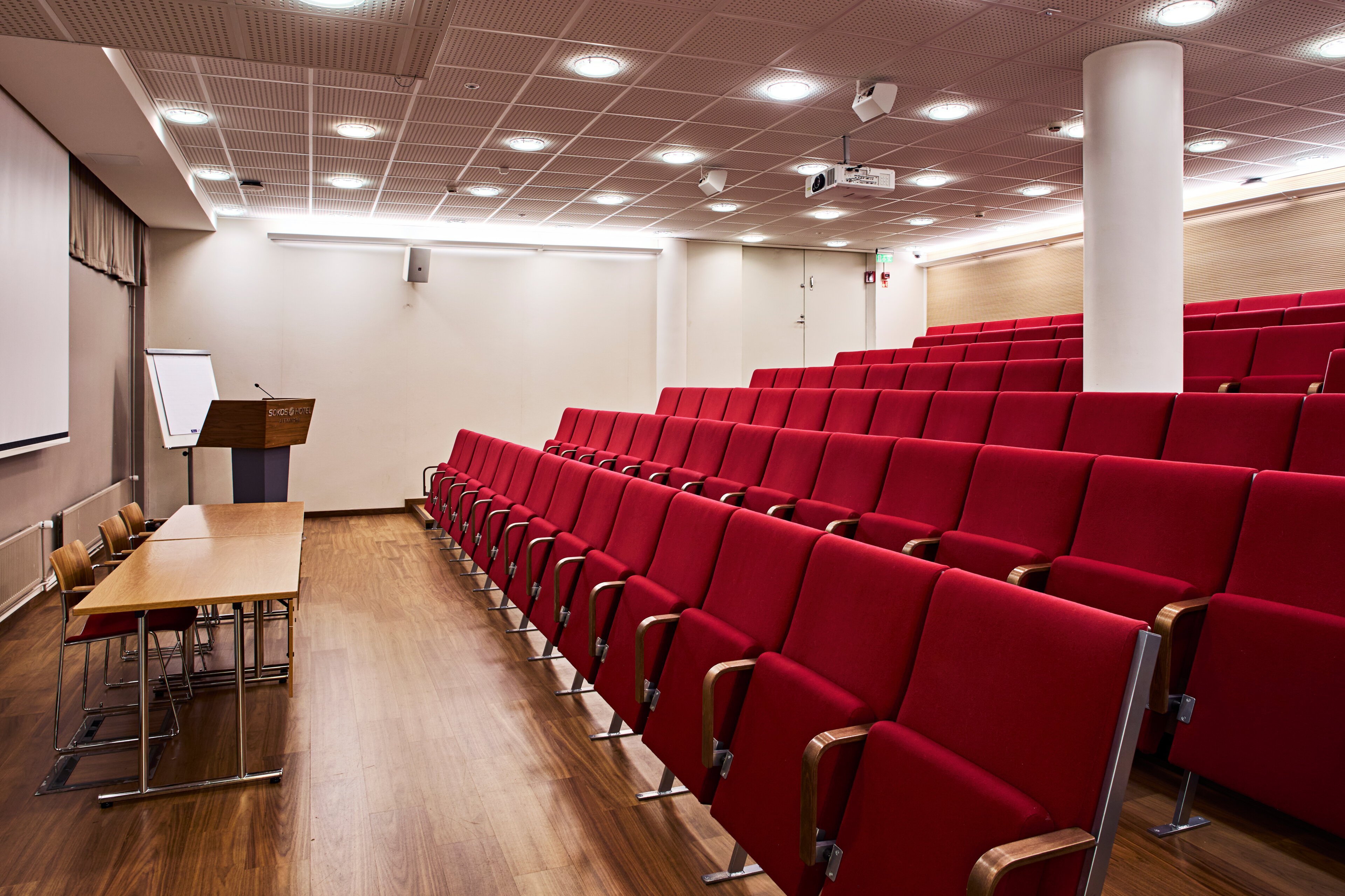 Auditorio punaisilla penkkiriveillä, etuosassa puinen puhujapöytä ja puhujakoroke. Puulattia ja valkoinen kattovalaistus.