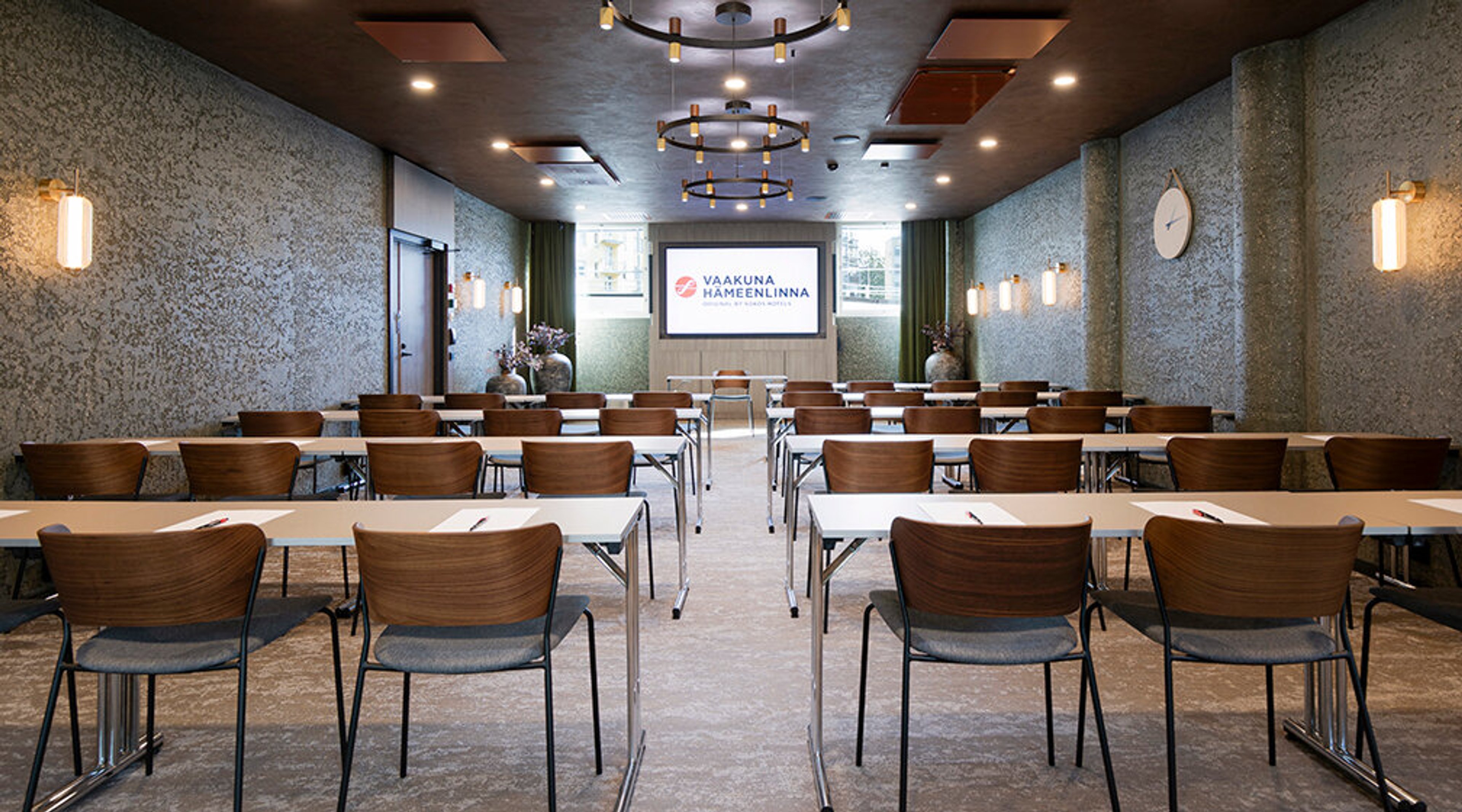 Modern conference room with brown chairs and white tables arranged in rows facing a presentation screen. Decorative light fixtures on the ceiling.