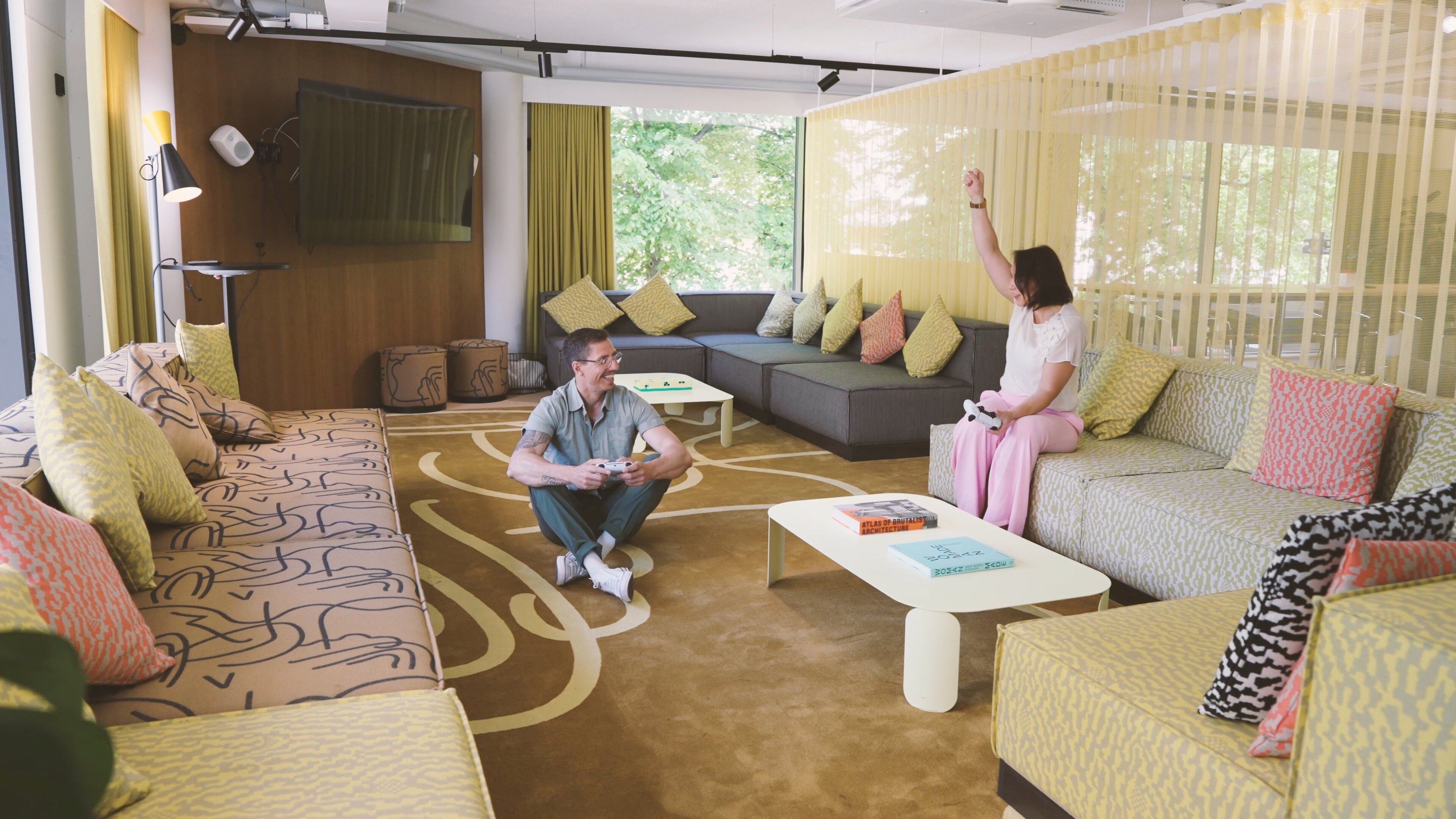 Modern lounge area with multiple sofas and colorful cushions. Spacious room with large windows, patterned carpet and two people playing video games.