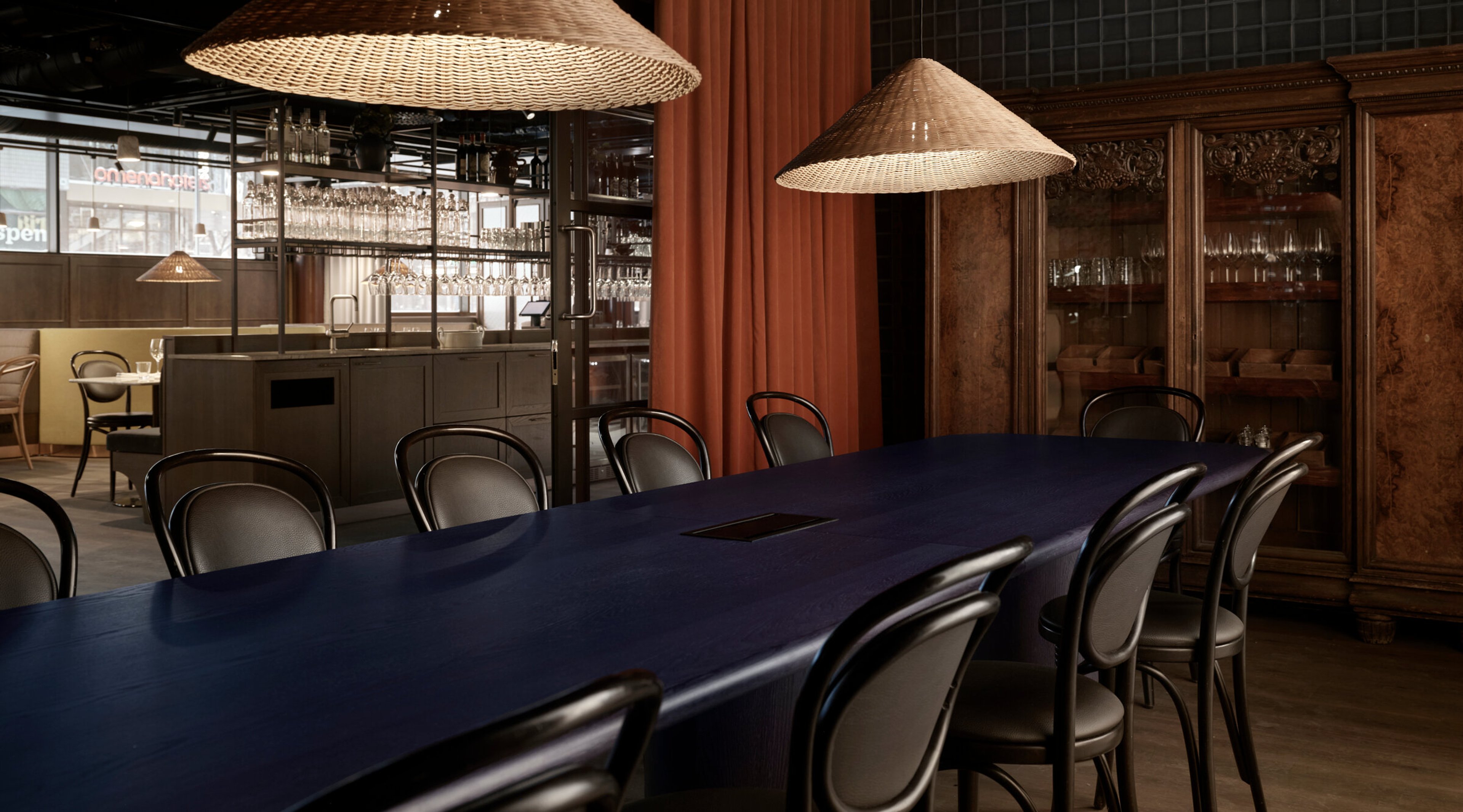 Restaurant interior with a long blue table and chairs. Wicker pendant lamps hang above, with a bar counter and red curtain in the background.