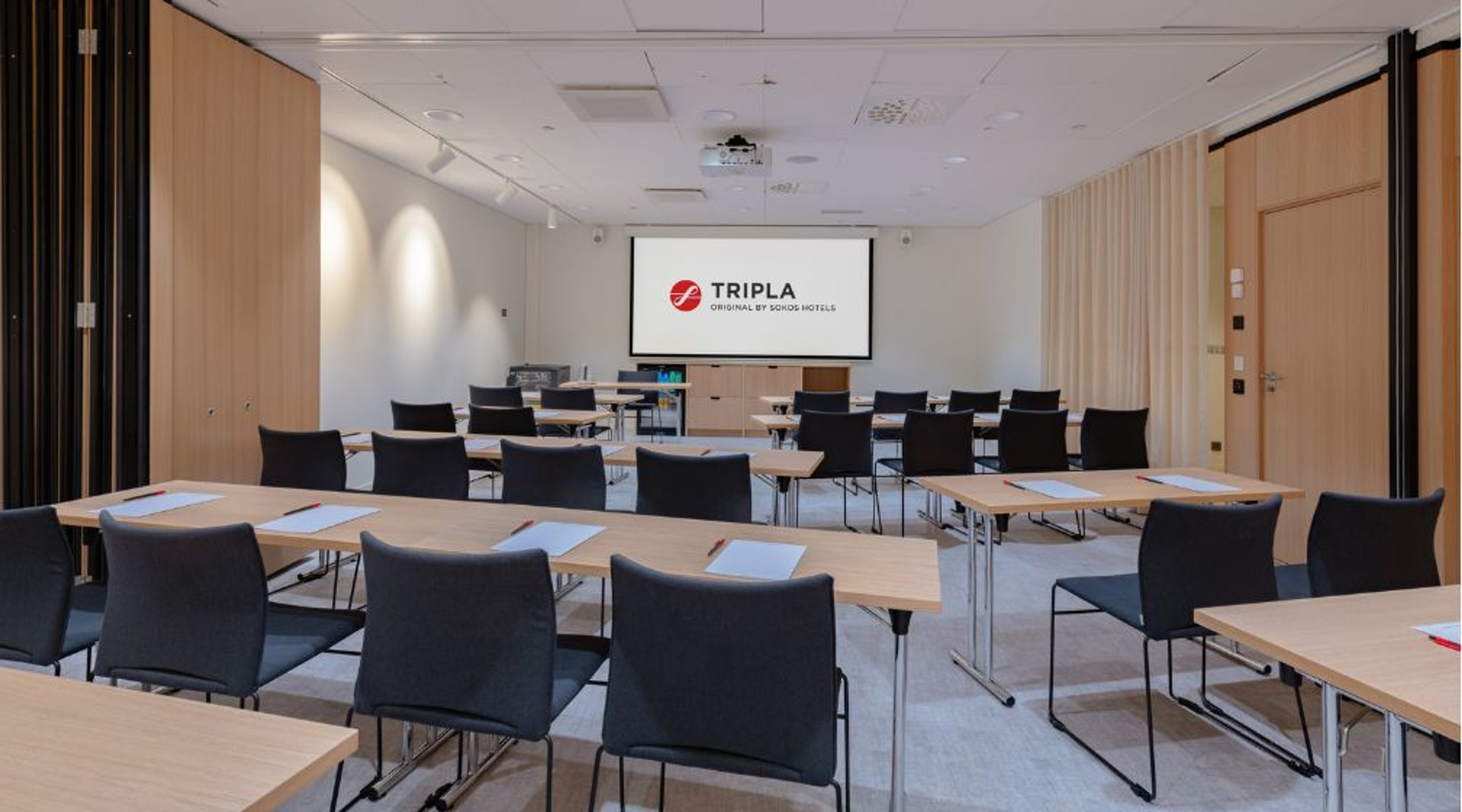 Moderni kokoustila vaaleilla puupaneeleilla, mustilla tuoleilla ja puisilla pöydillä. Edessä valkokangas projektorille. Tila on tasalattiallinen.