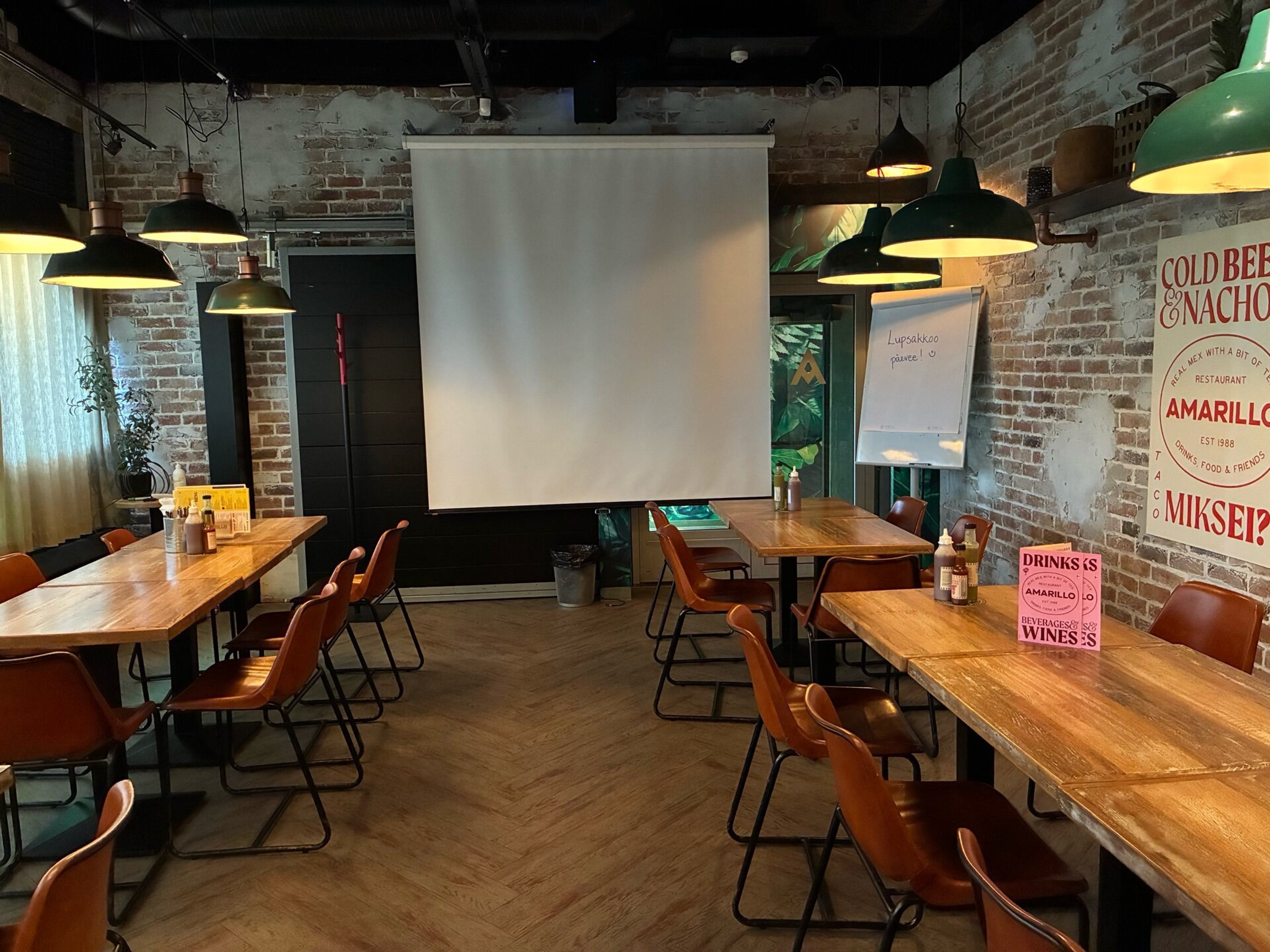 Ravintilasta konferenssikäyttöön muunnettu tila, jossa on tiilikiviseinät, puiset pöydät ruskeilla tuoleilla ja valkokangas. Lattia on puiset parkettipalkit.