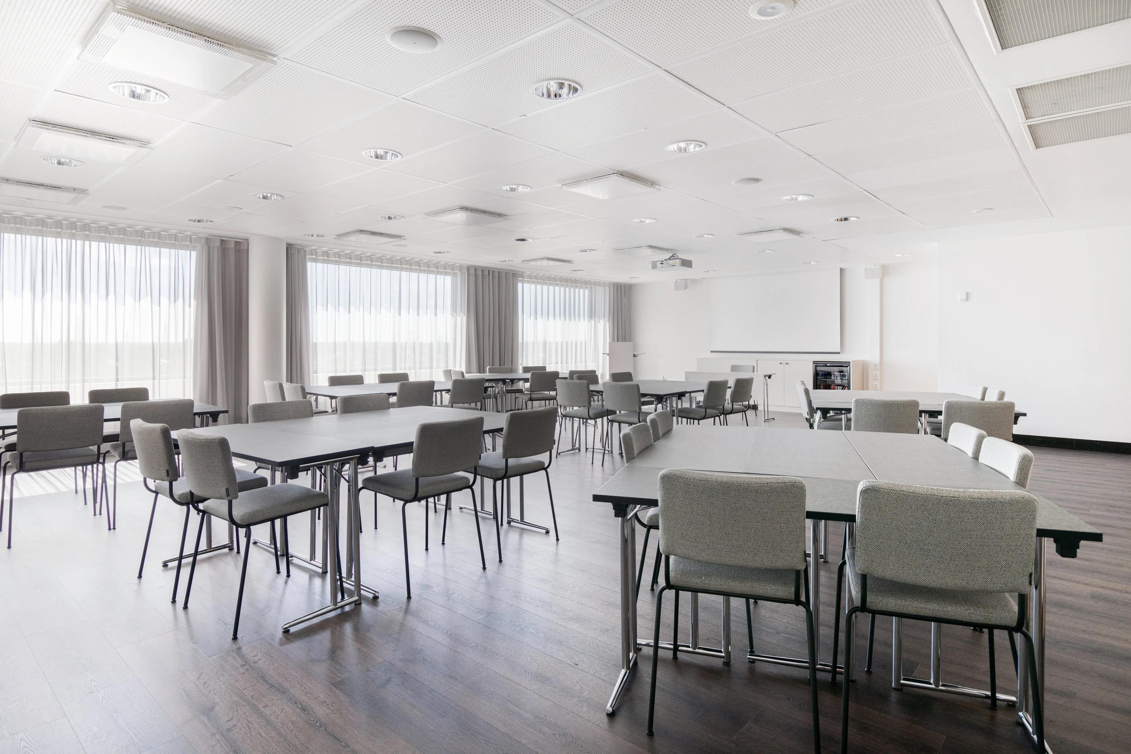 Bright meeting room with grey chairs and rectangular tables. Smooth wooden floor and large windows. Projection screen on wall for presentations.