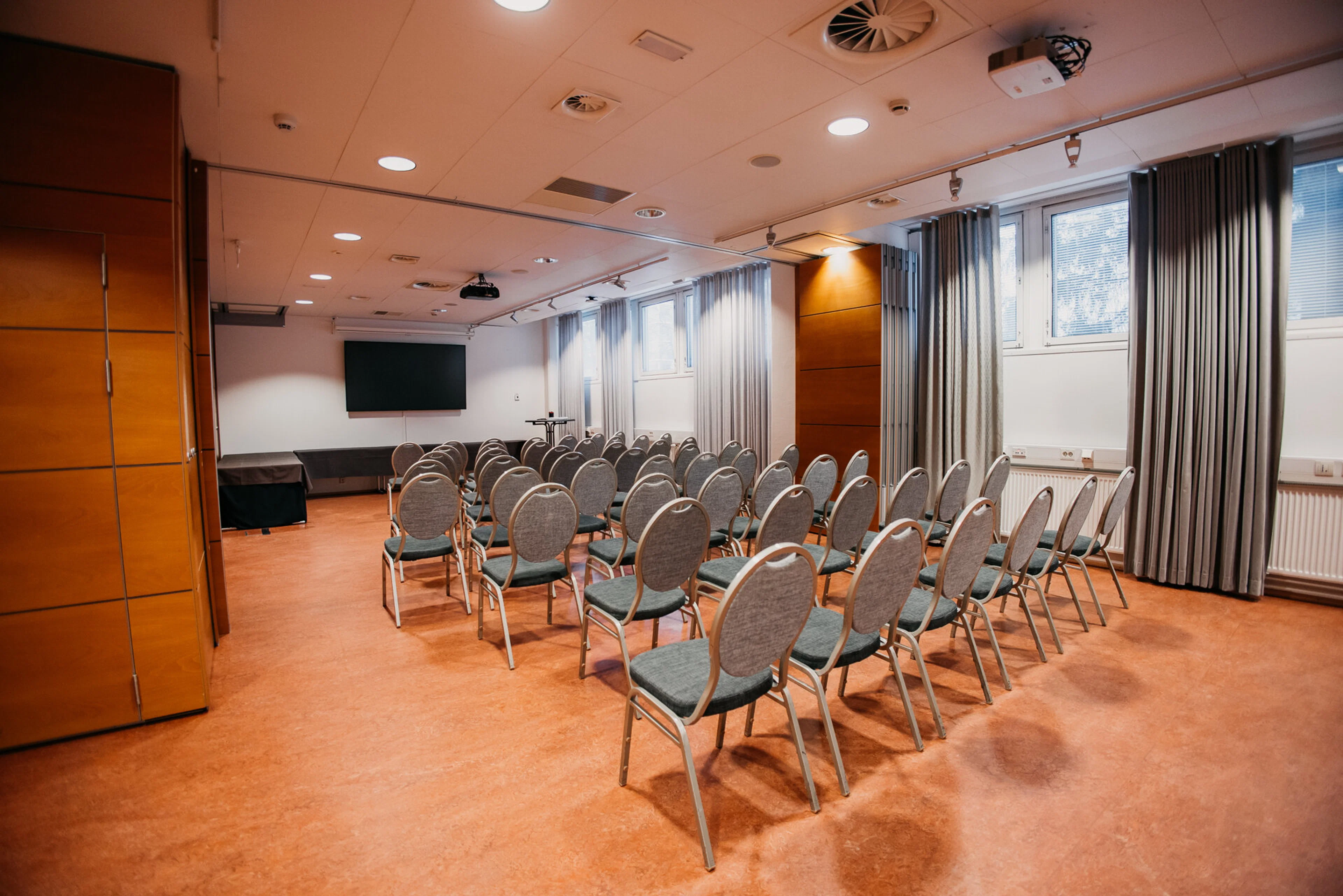 Konferensrum med grå stolar i rader, projektor, skärm och träpanelerade väggar. Plant golv och god belysning.