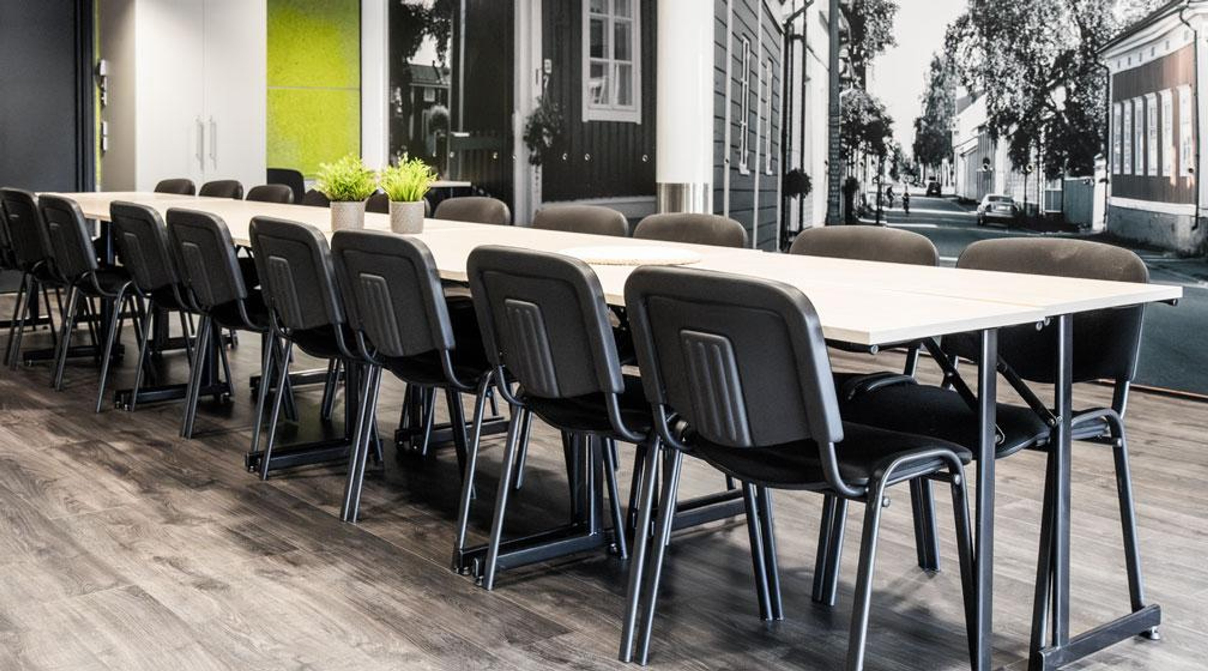 Conference room with a long white table surrounded by black chairs. Wall featuring urban scenery and green design element in background. Wooden floor.