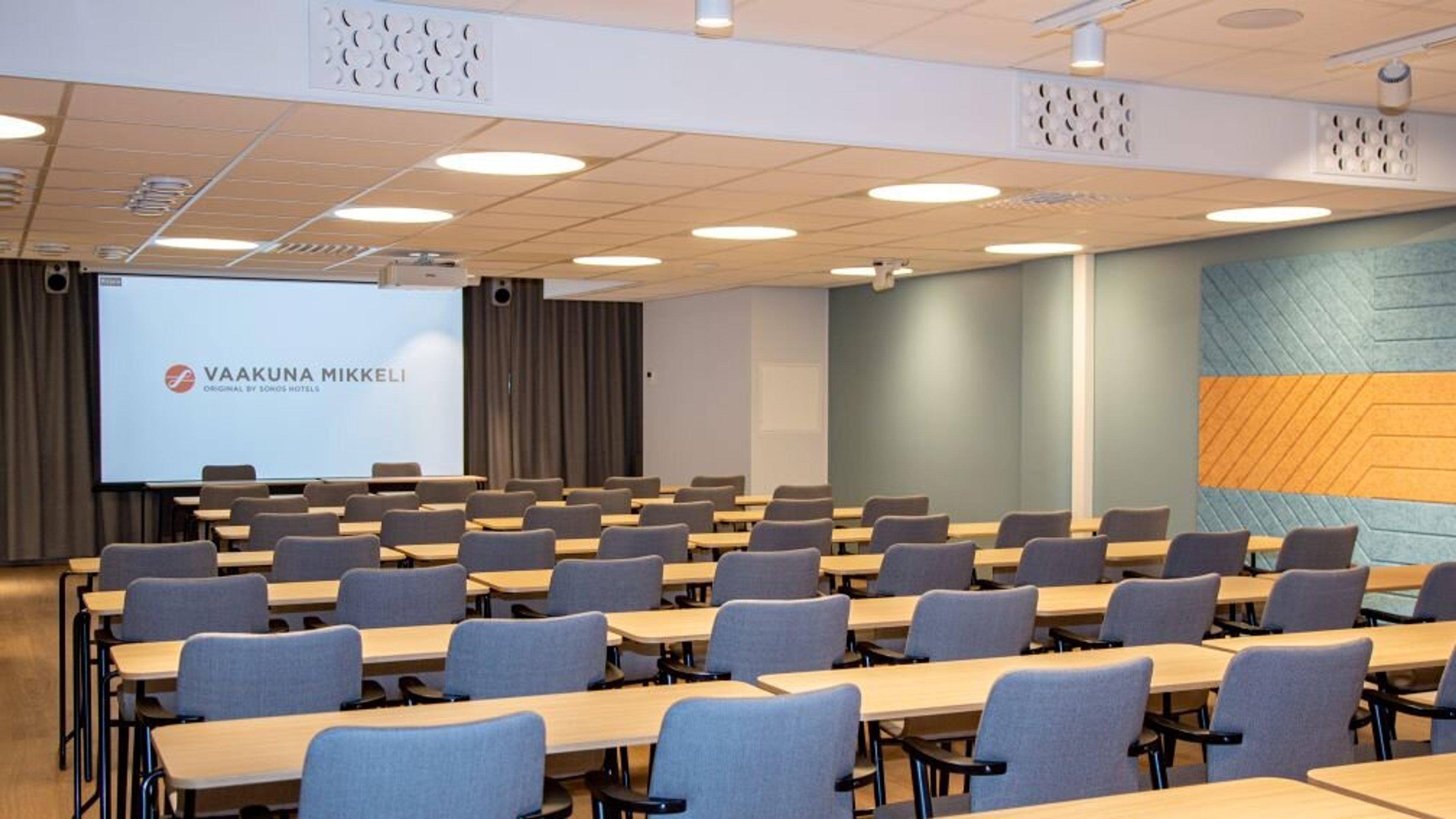 Föreläsningssal med rader av bord och blå stolar, projektorduk framme. Färgglada akustikpaneler på väggen.