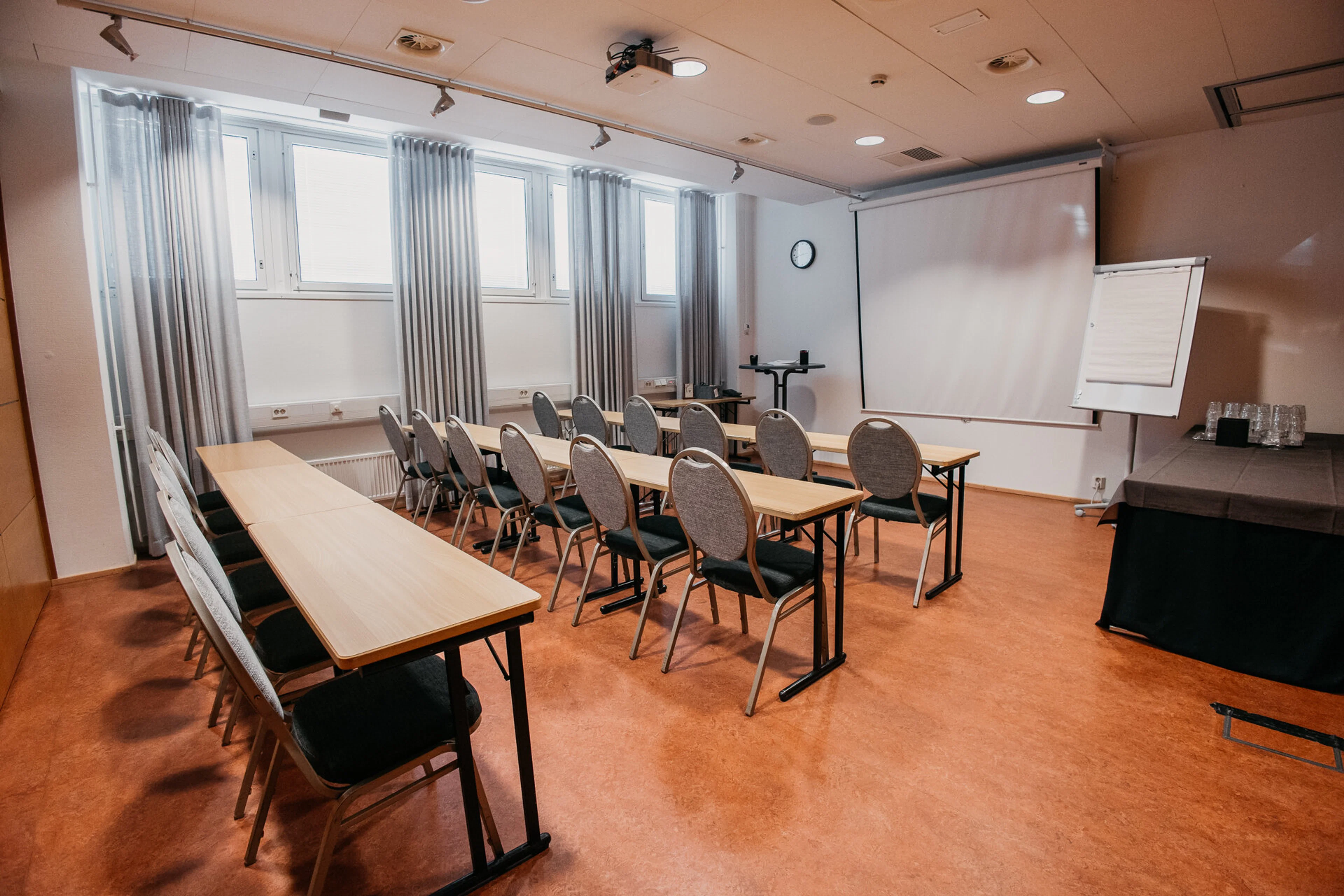 Litet mötesrum med bord i klassrumsformat, projektorduk framme och fönster på sidoväggen. Jämnt hårt golv och gott om utrymme att röra sig.